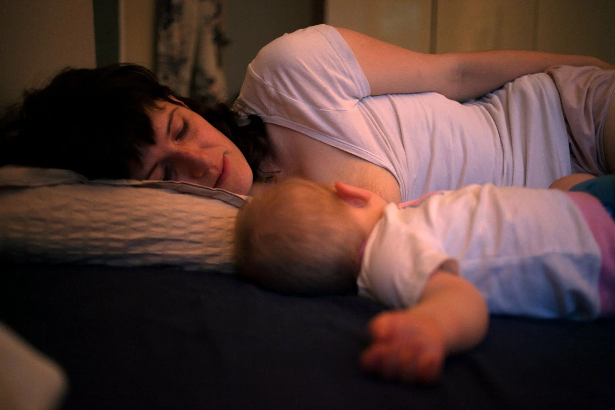 A woman with dark hair lying on her side on a bed, looking at a baby with blonde hair who is lying on their back, both appearing to be in a tender moment.