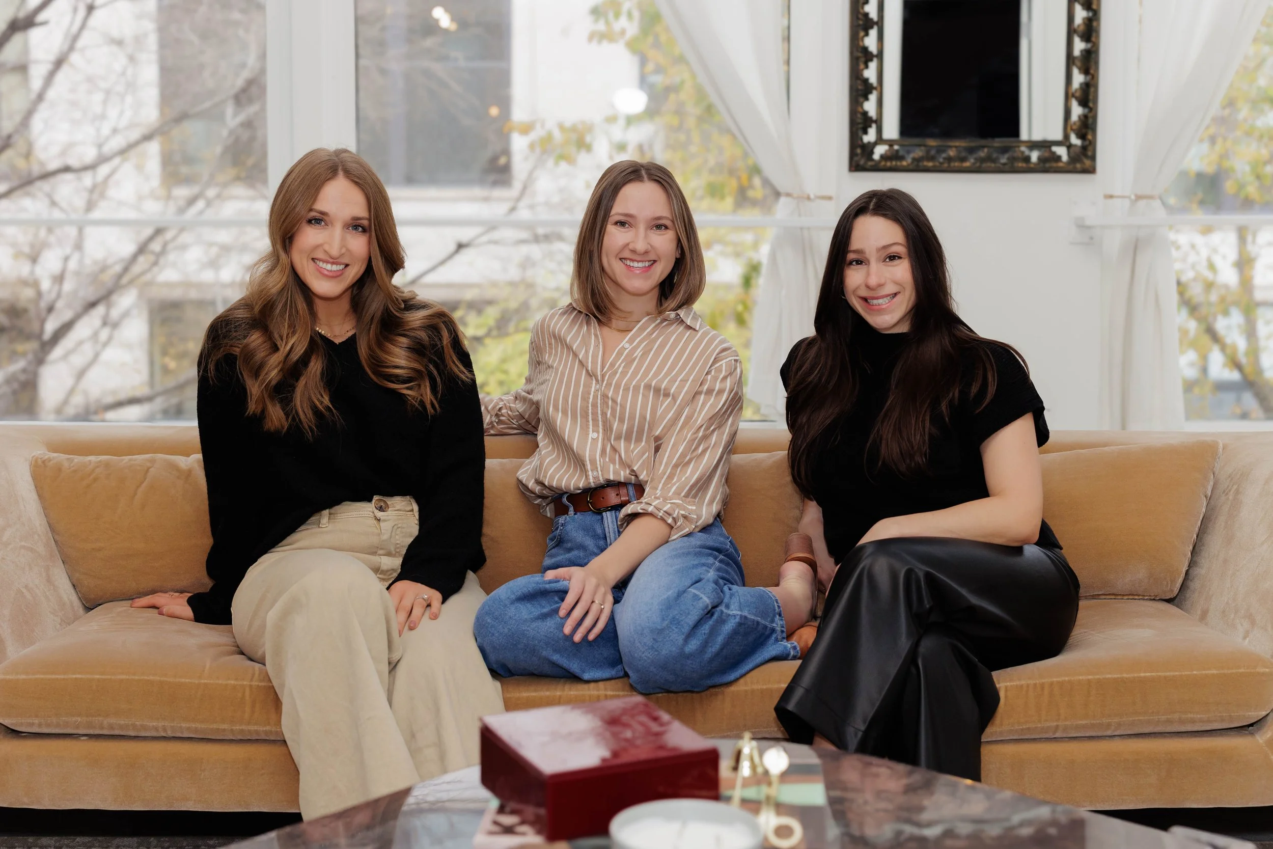 Three women sitting on a beige couch in front of large windows with white curtains, smiling at the camera.