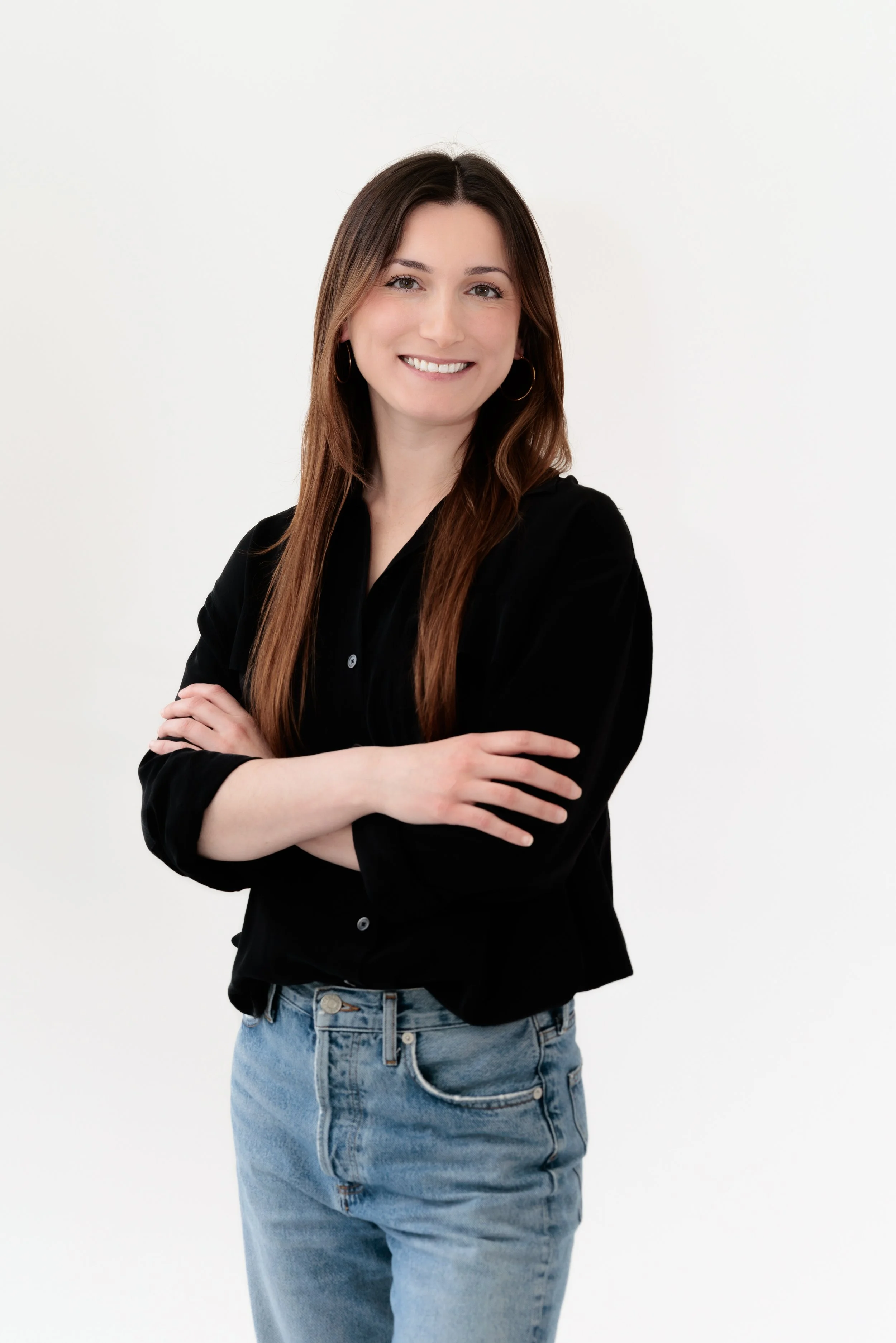 A smiling woman with long brown hair wearing a black button-up shirt, standing against a light grey background.