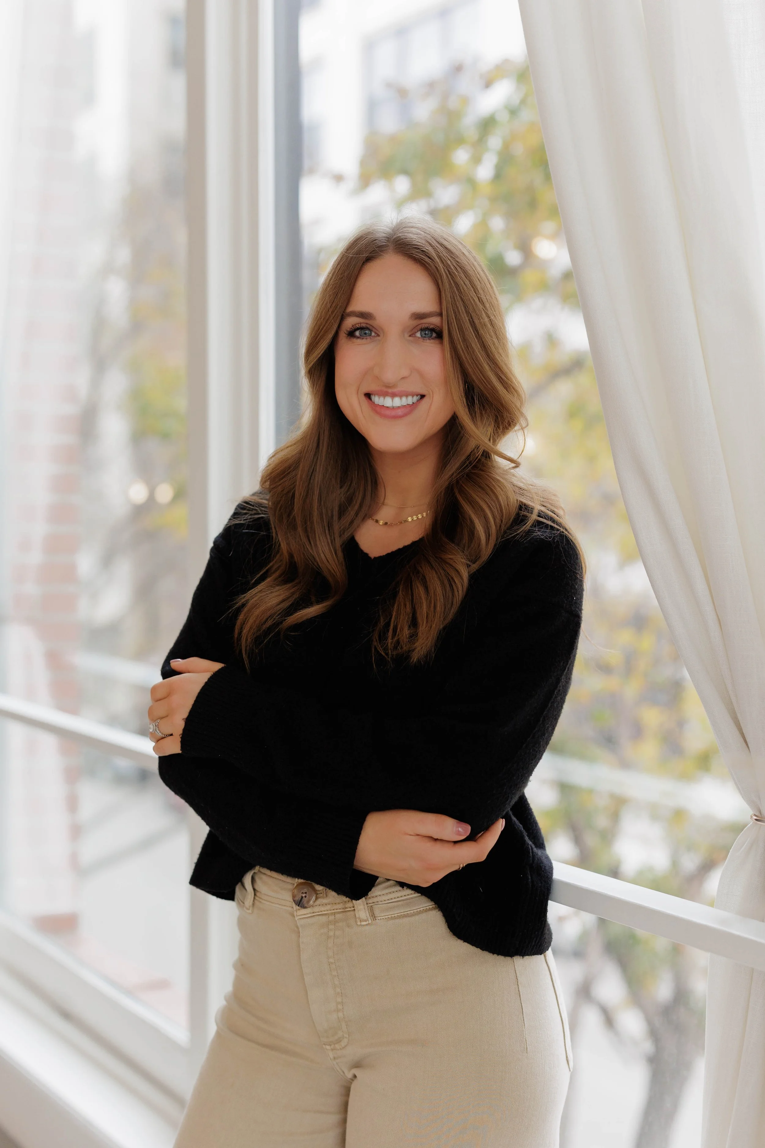 Mollie Bass, LMFT, in black sweater and beige pants smiling while standing by a window with natural light and a view of trees outside.