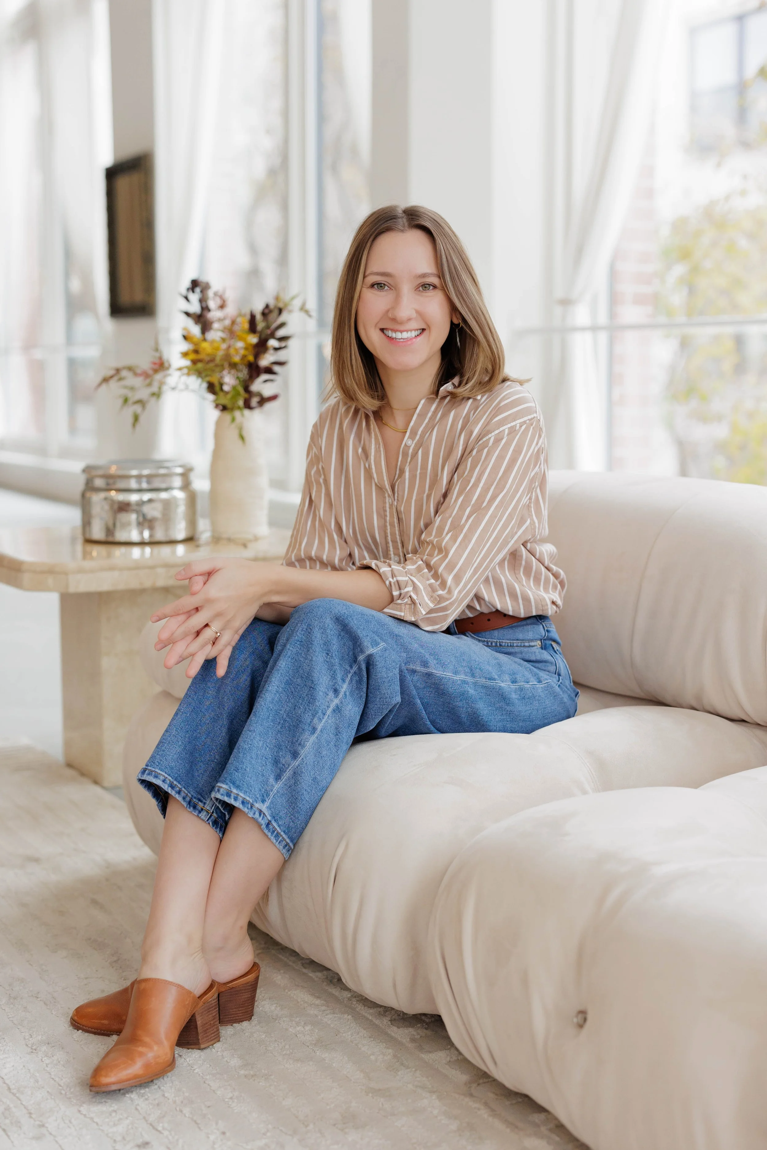 Alana Stramowski, LPC, sitting on a beige couch in a bright room with large windows. She is wearing a striped shirt, blue jeans, and brown heeled shoes. A table with a vase of flowers and a metal container is in the background.