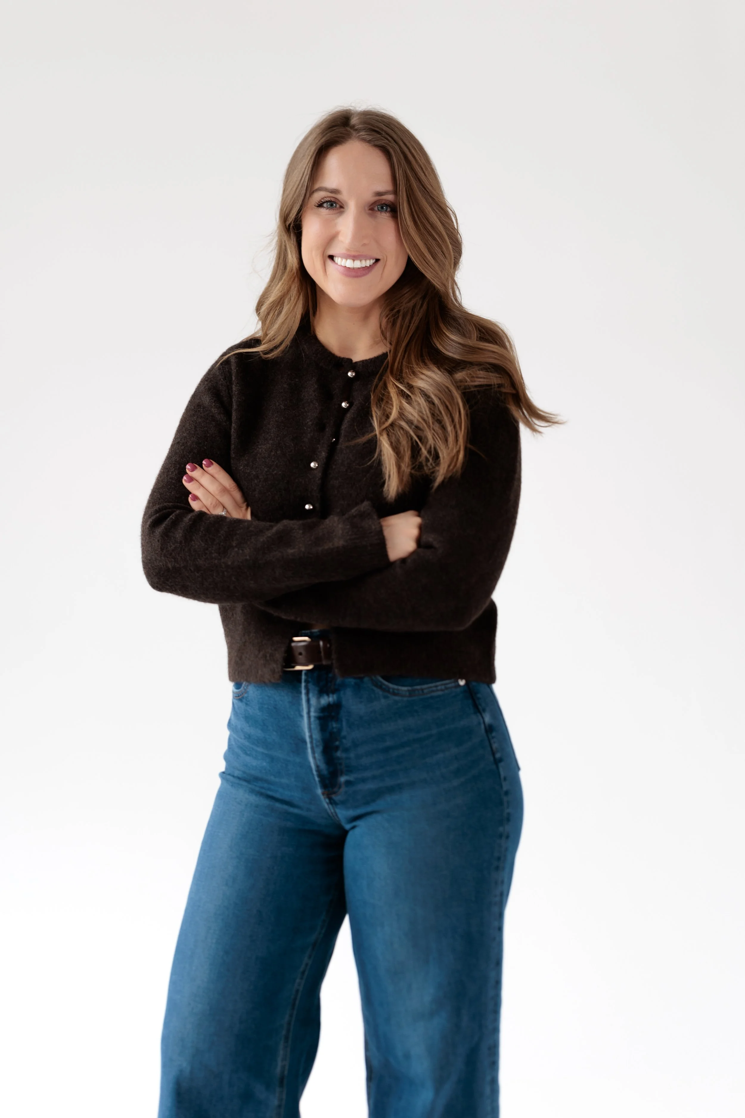 A smiling woman with long, wavy brown hair wearing a brown sweater, sitting against a plain light  background with her hands in her lap..