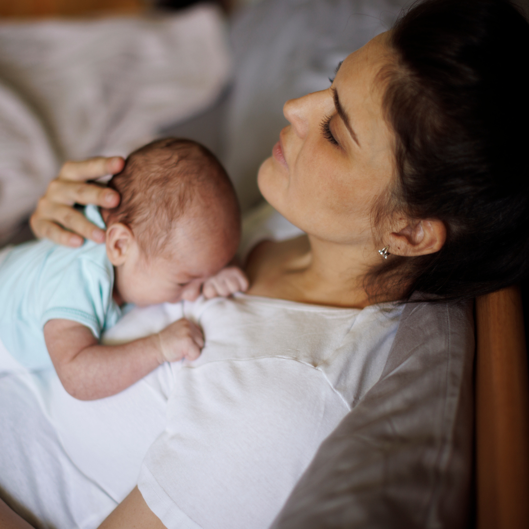 Parent lying in bed, holding a newborn on their chest, staring off into the distance—conveying the mental and emotional fog of postpartum adjustment.
