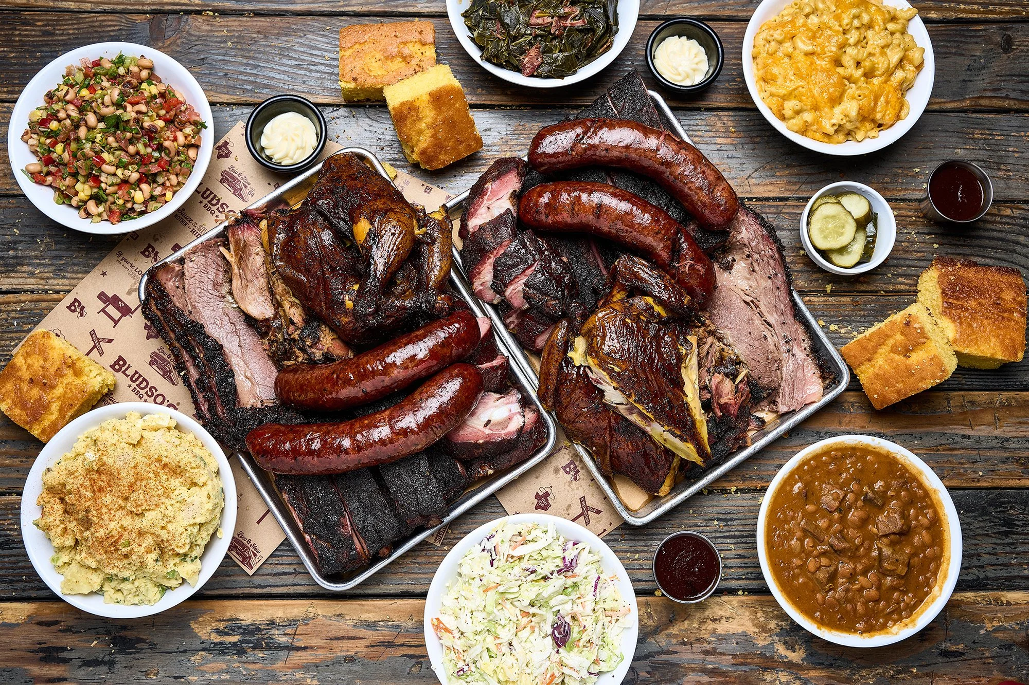 Two Bludso's BBQ trays overflowing with smoked meats surrounded by Bludso's side orders on a wo
