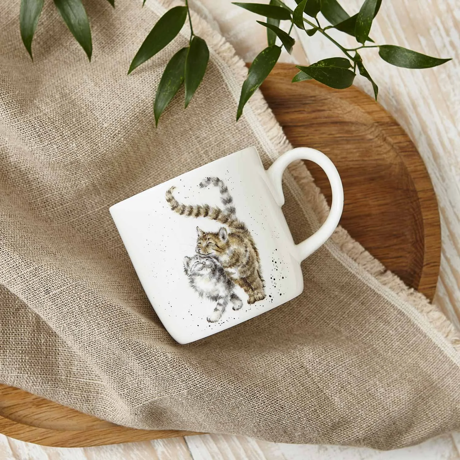 White mug with cats on the outside laying on a linen, each on a table.