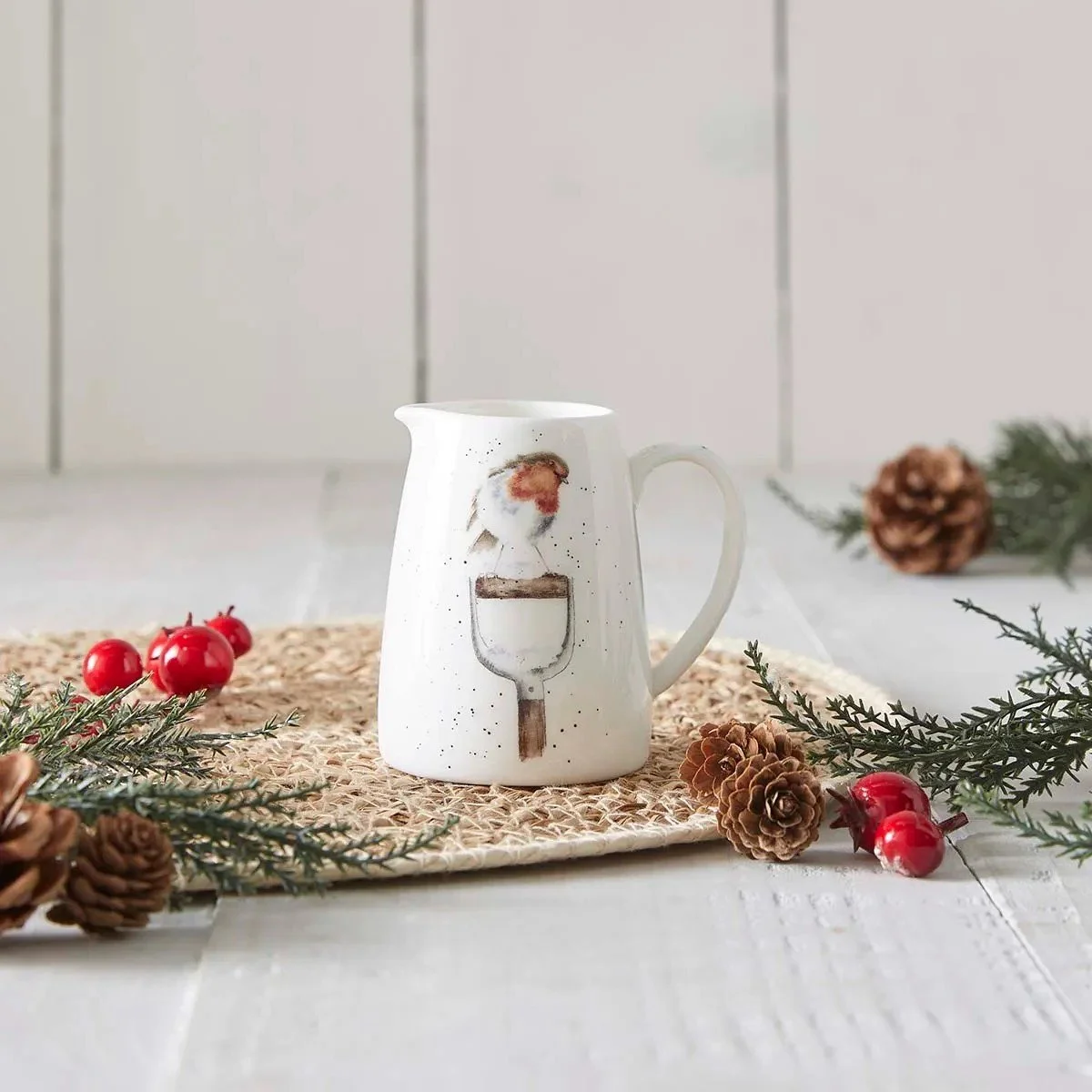 jug with robin on table with greenery, cranberries and pinecones.