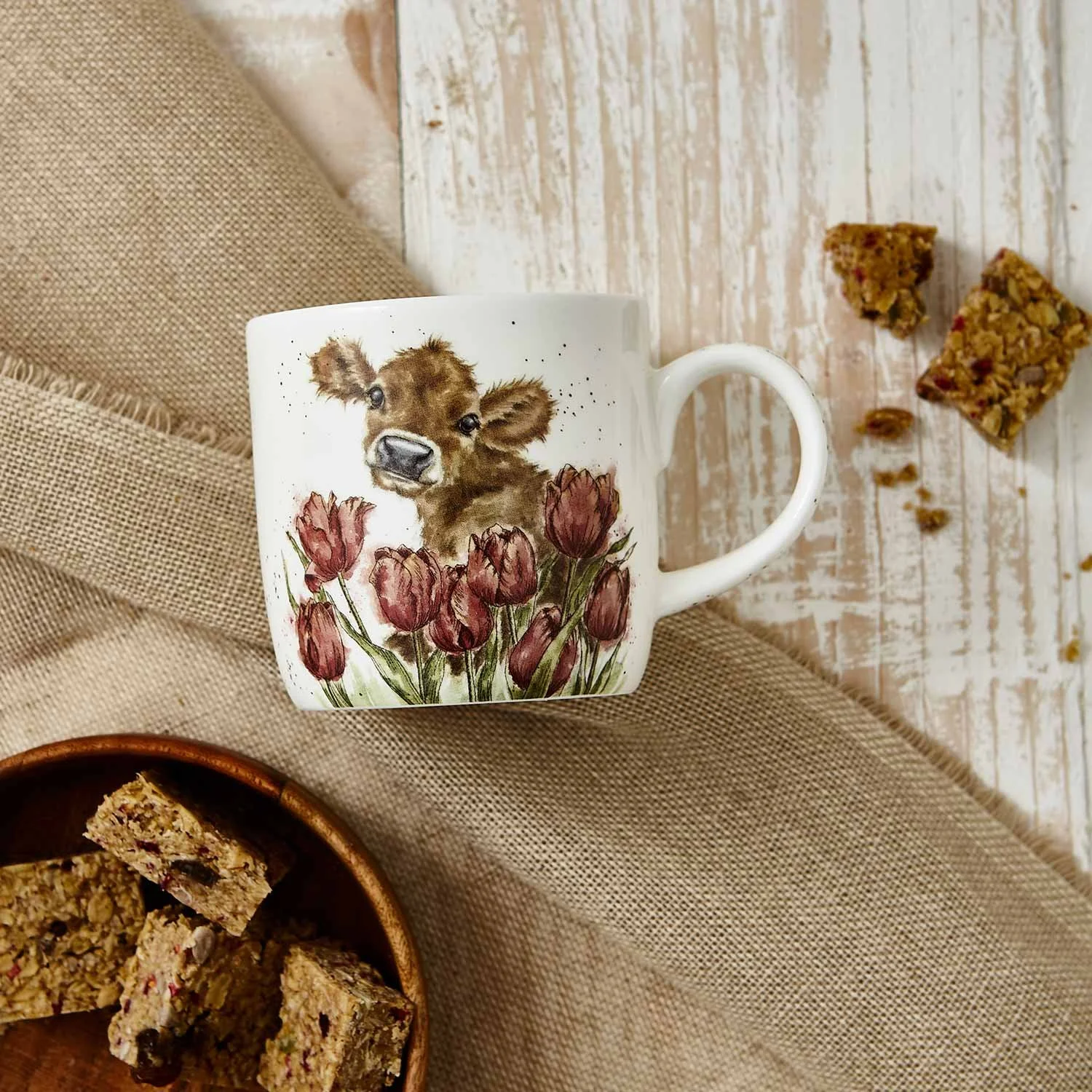 White mug on table that features brown cow and pink tulips.