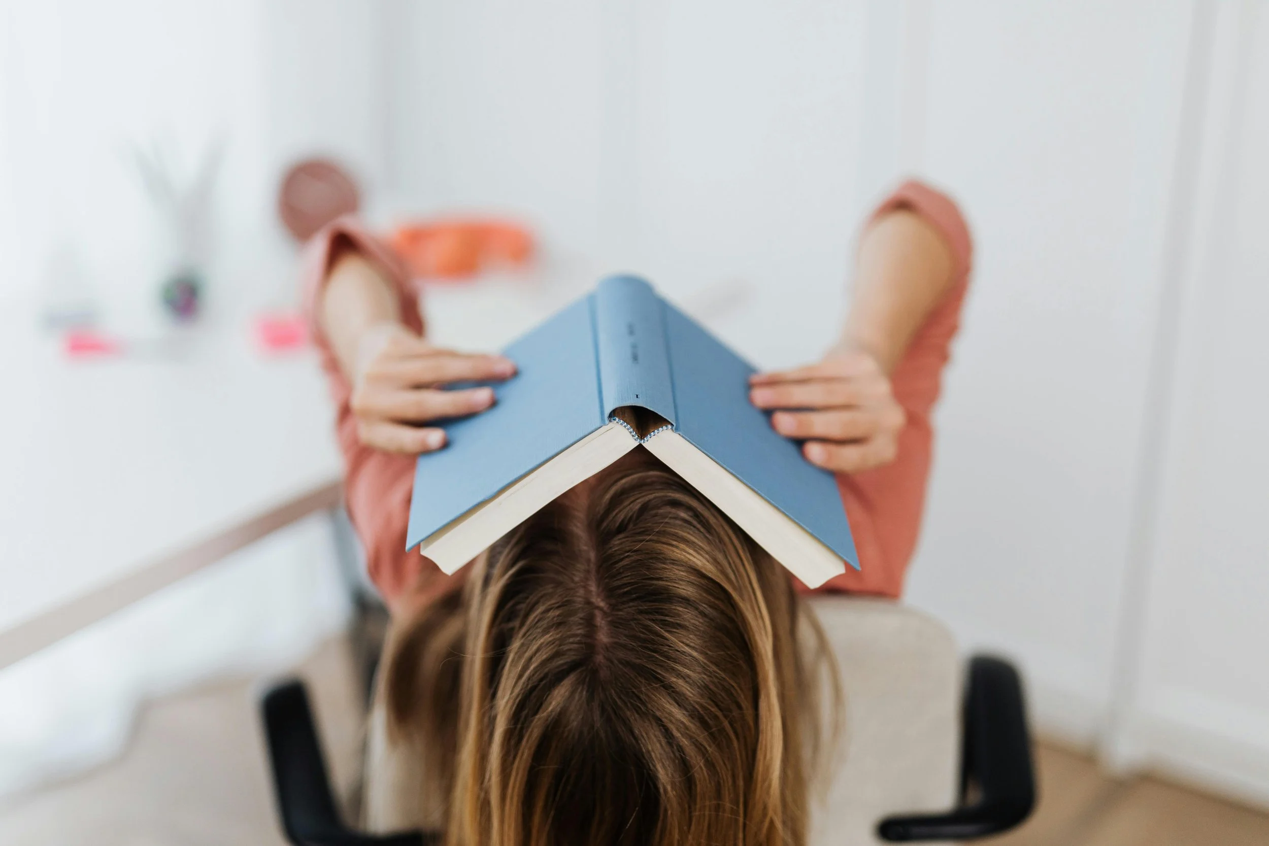 Woman sitting at a desk with a book covering her face.