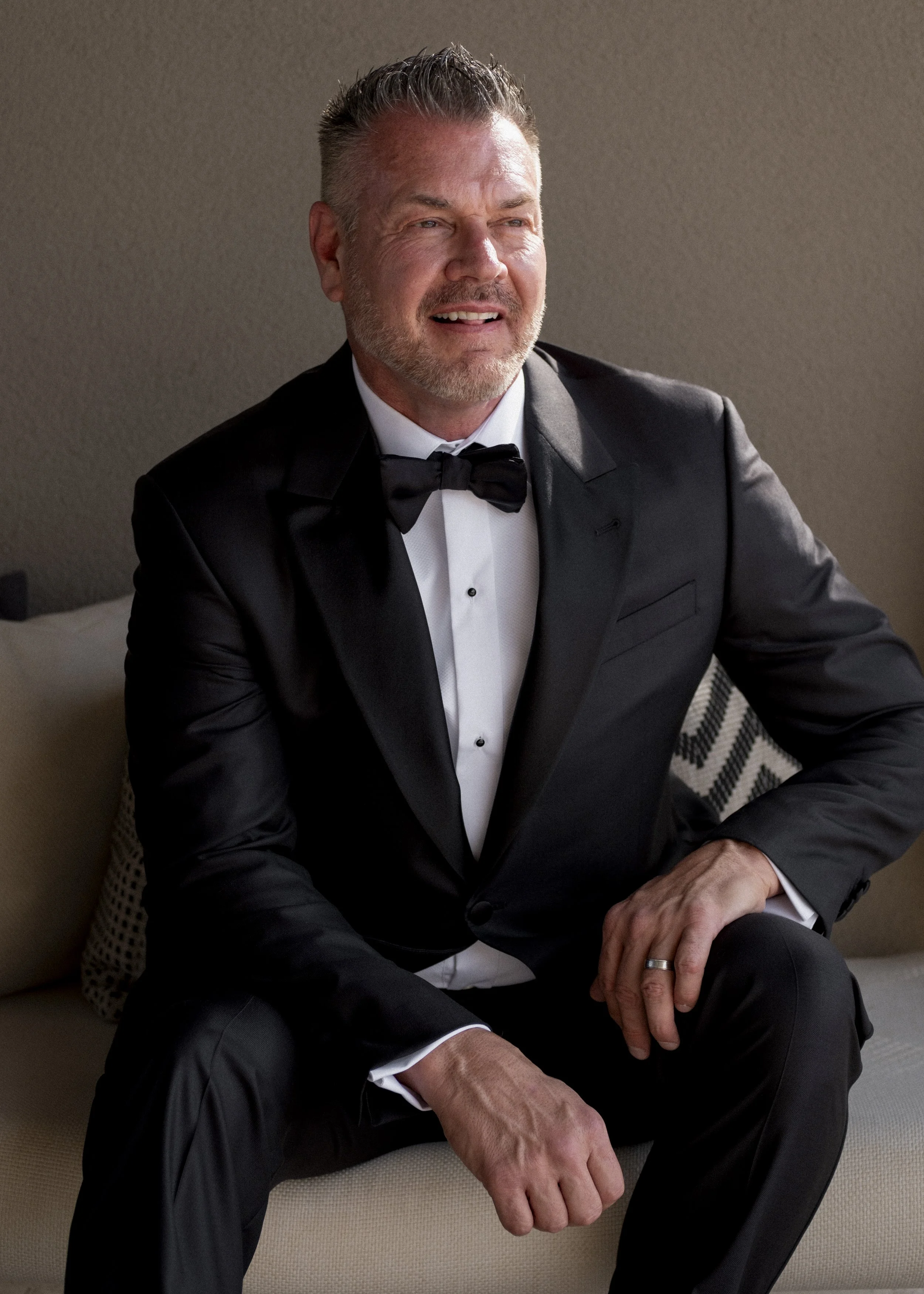 A man wearing a tuxedo and bow tie sitting on a beige couch, smiling, with a neutral wall background.