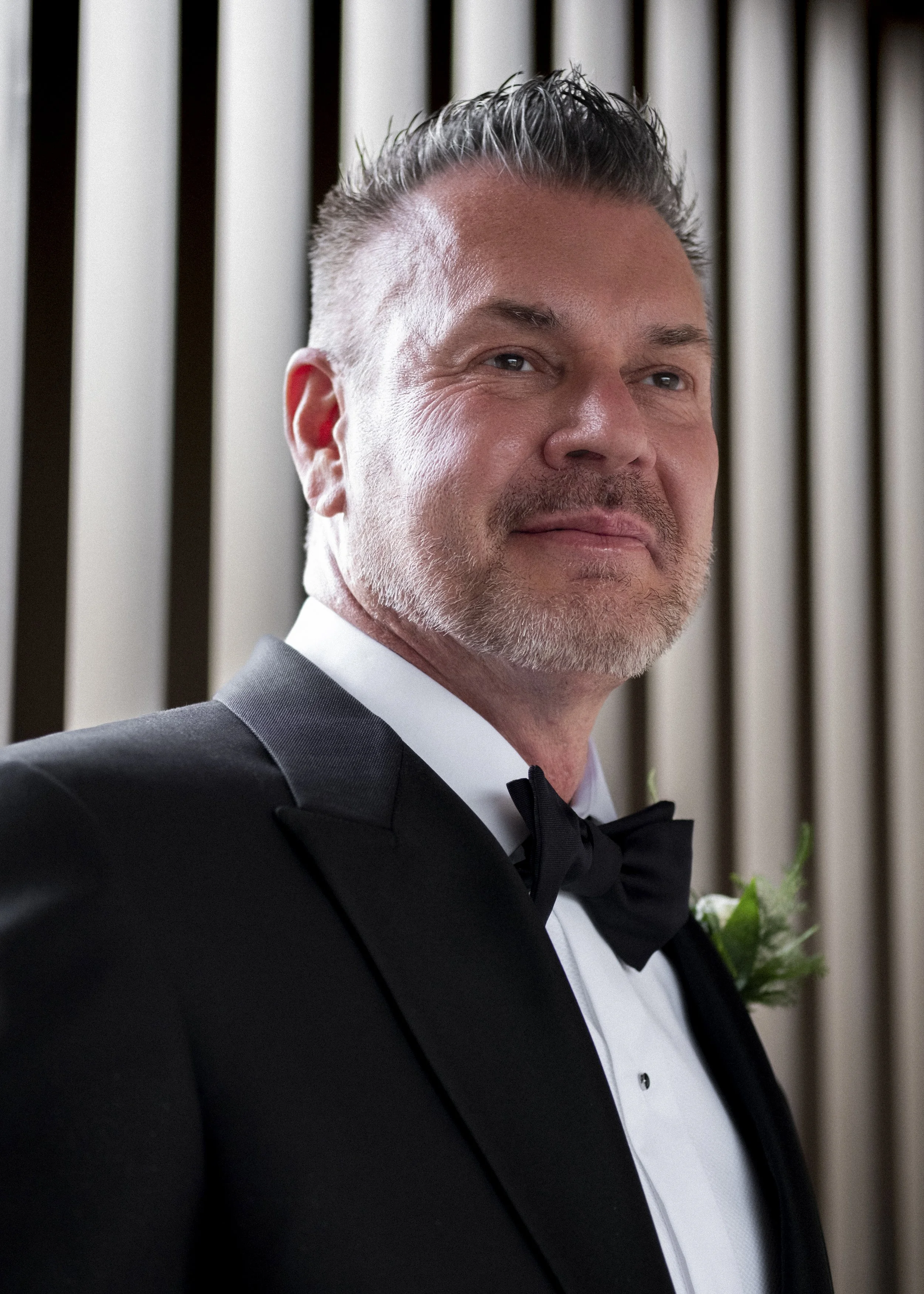 A man in a tuxedo with a bowtie, smiling and standing indoors with vertical blinds in the background.