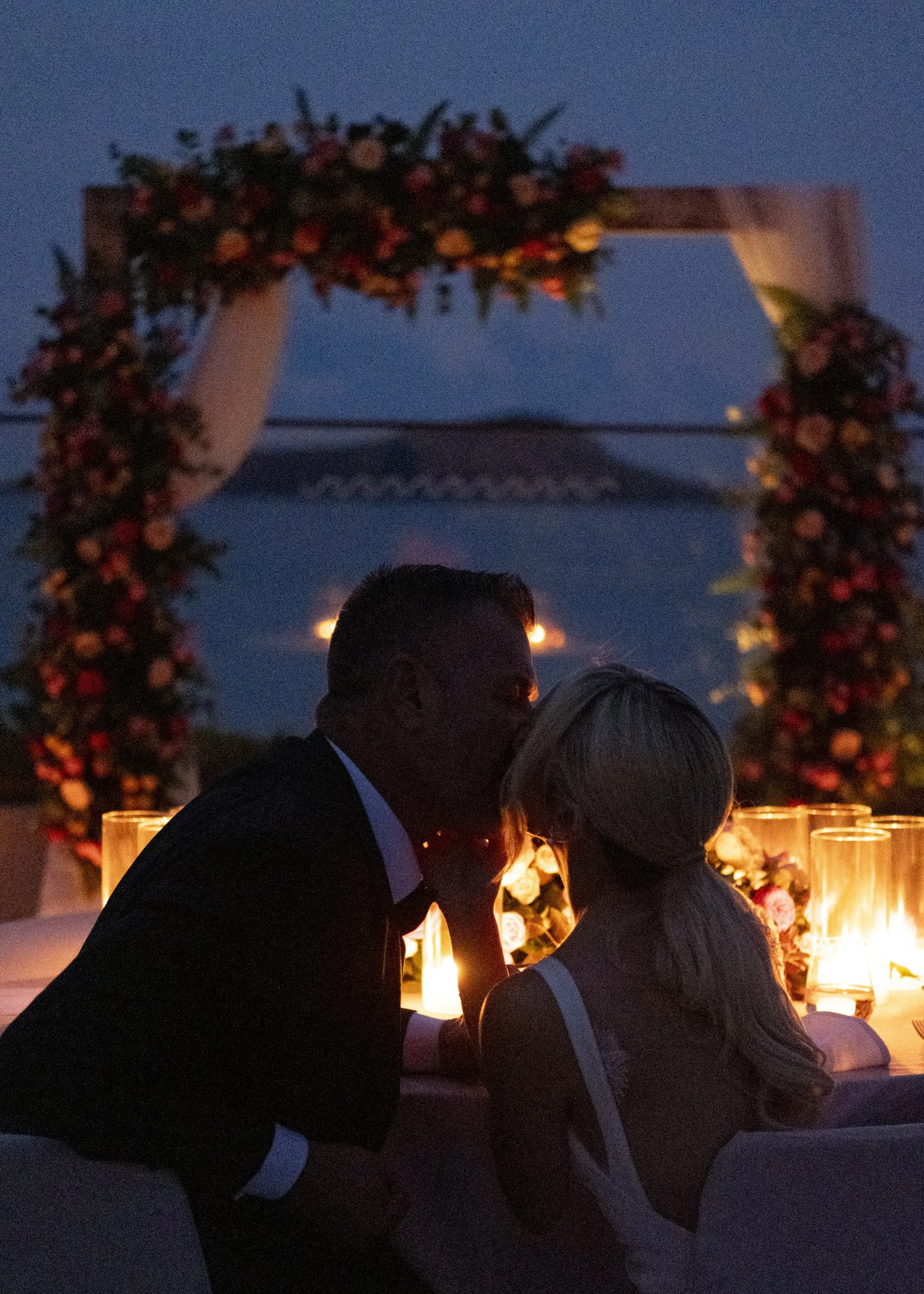A couple sharing a kiss at a romantic outdoor dinner during the evening, with a floral arch and candlelit tables in the background.