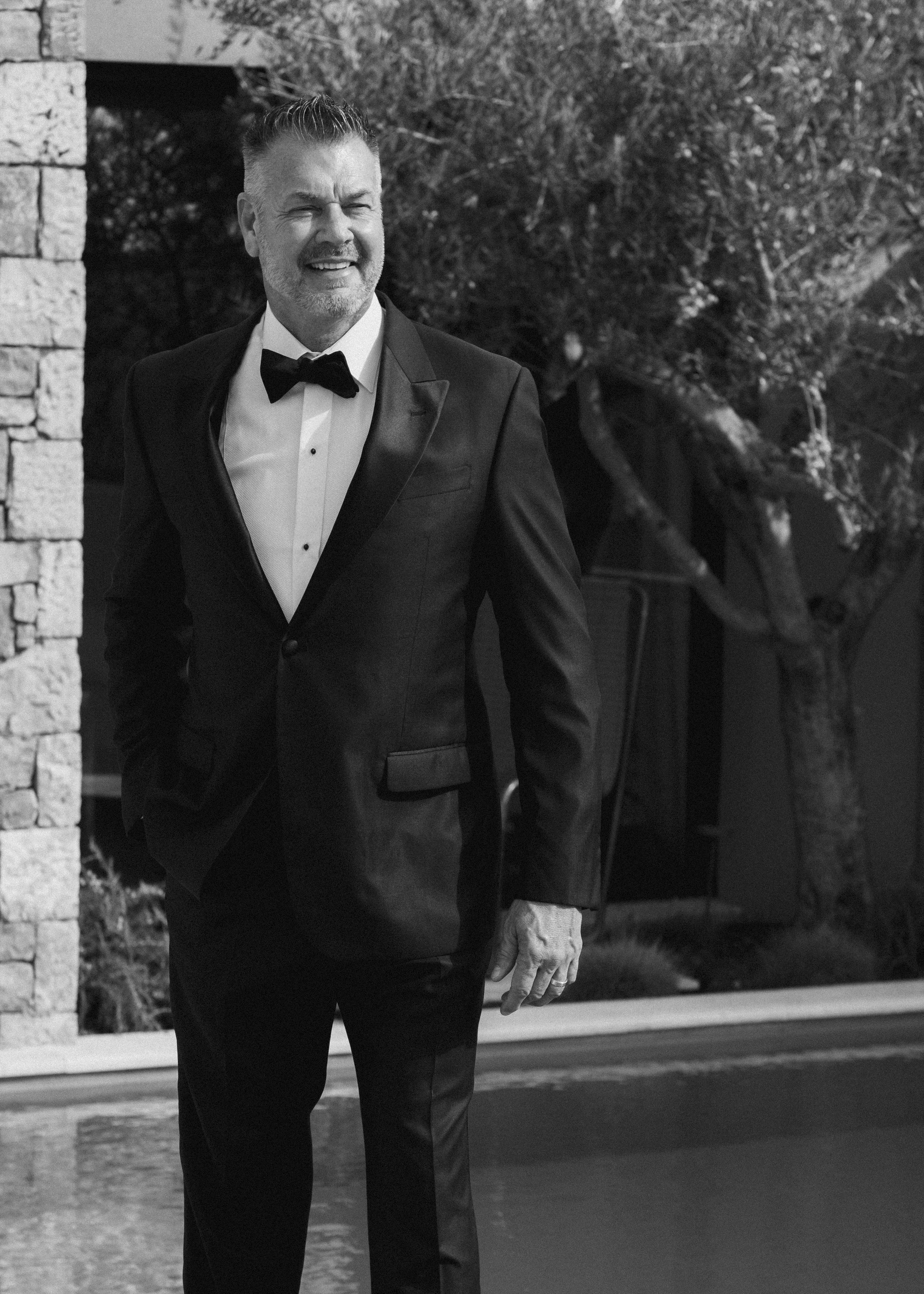 A man in a tuxedo with a bow tie standing outdoors near a stone wall and trees, smiling with a relaxed pose.