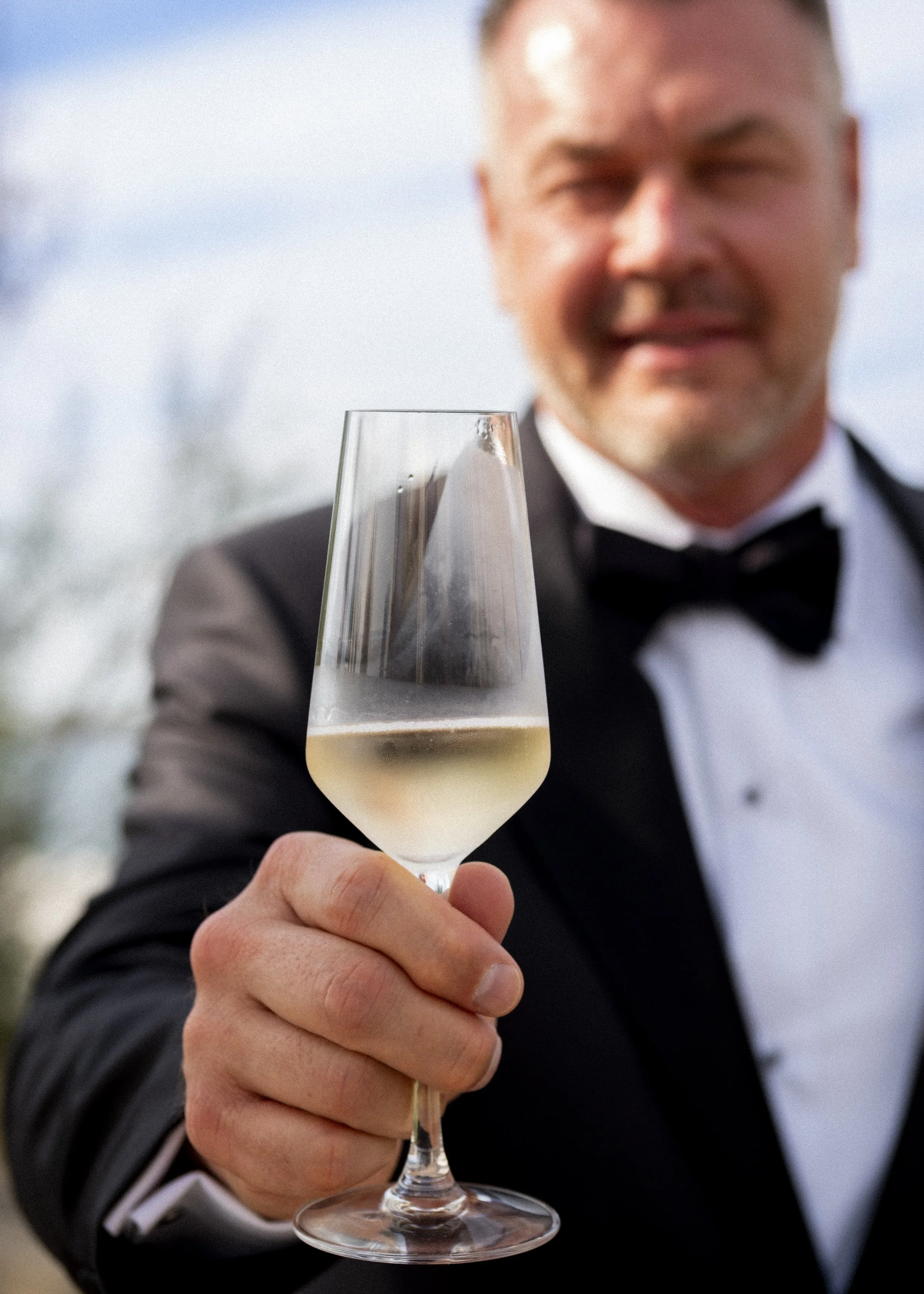 A man in a tuxedo holding a glass of champagne, with a blurred background of trees and sky.