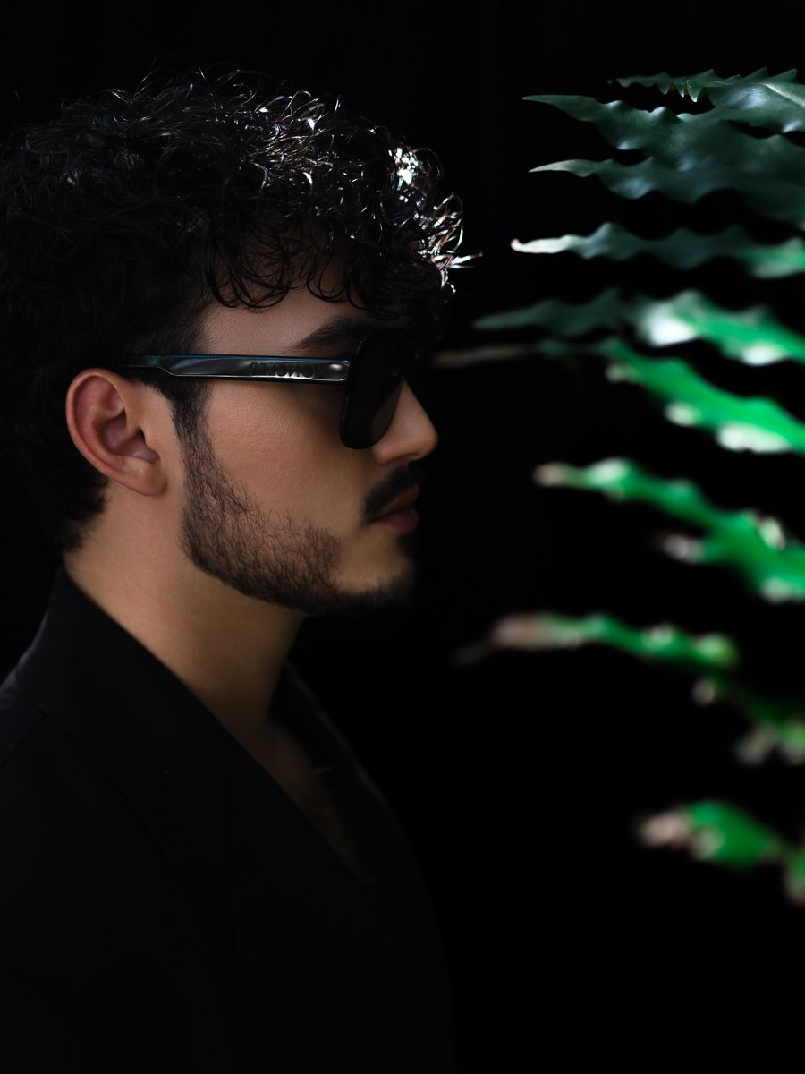 Side profile of a man with curly hair and a beard wearing dark sunglasses against a black background with green foliage.