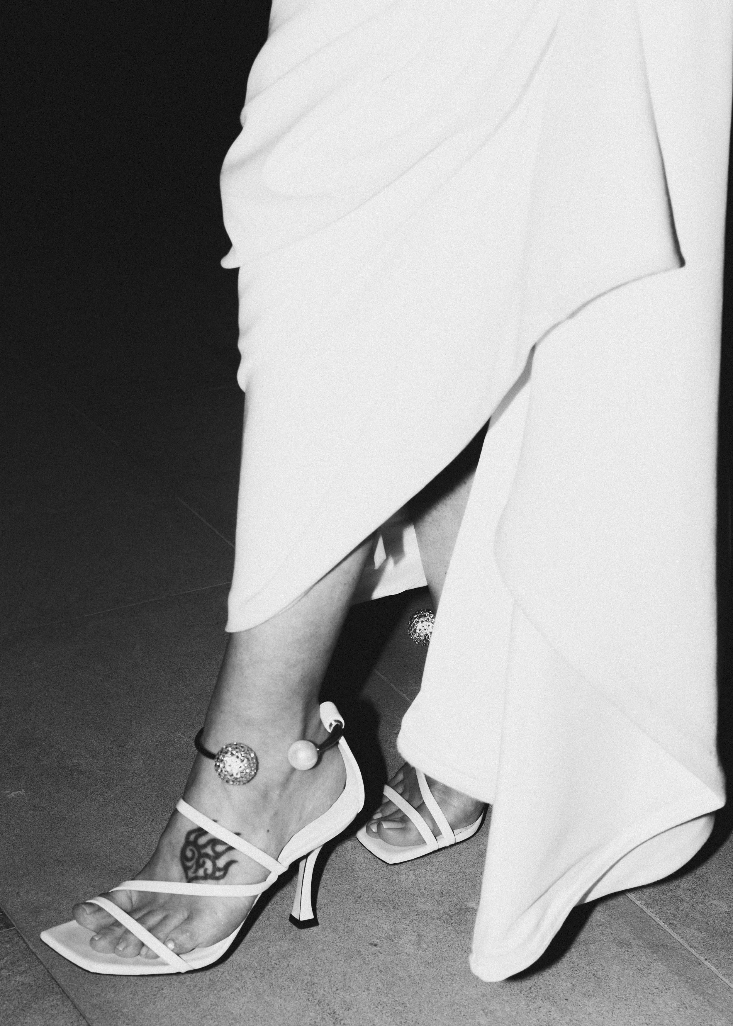 Black and white photo of a person wearing high-heeled sandals with an ankle bracelet, exposing a tattoo on the foot, and wearing a long skirt that is lifted to reveal the legs.