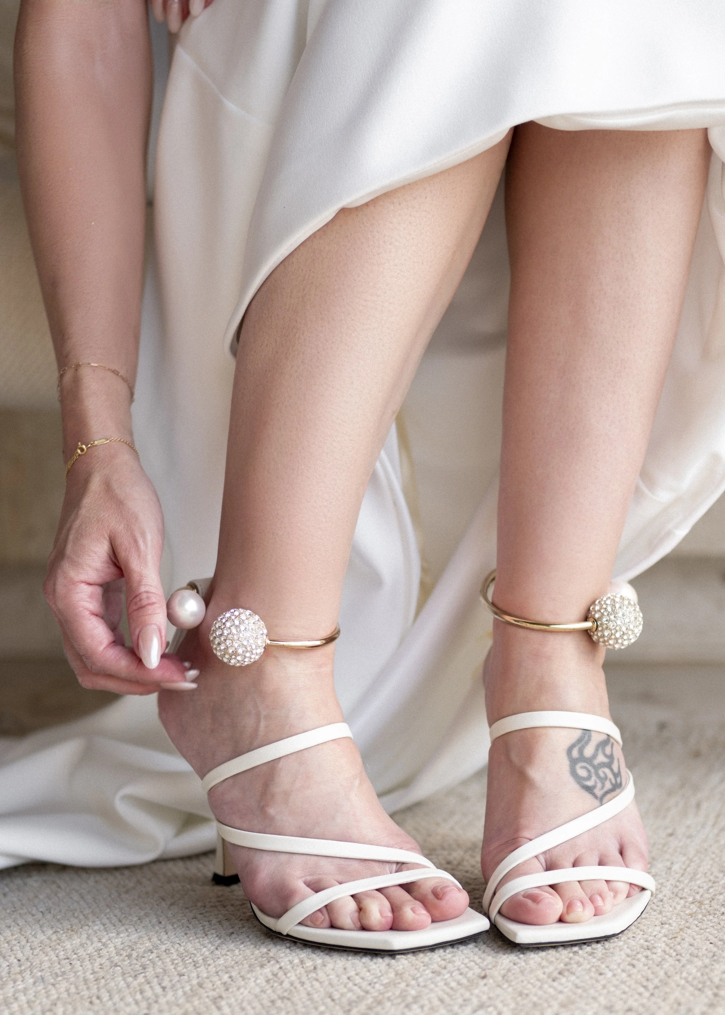 Close-up of a woman's legs and feet, wearing white high-heeled sandals, gold bracelet with decorative balls, and anklets with large pearl and rhinestone ornaments, with a tattoo visible on her ankle.