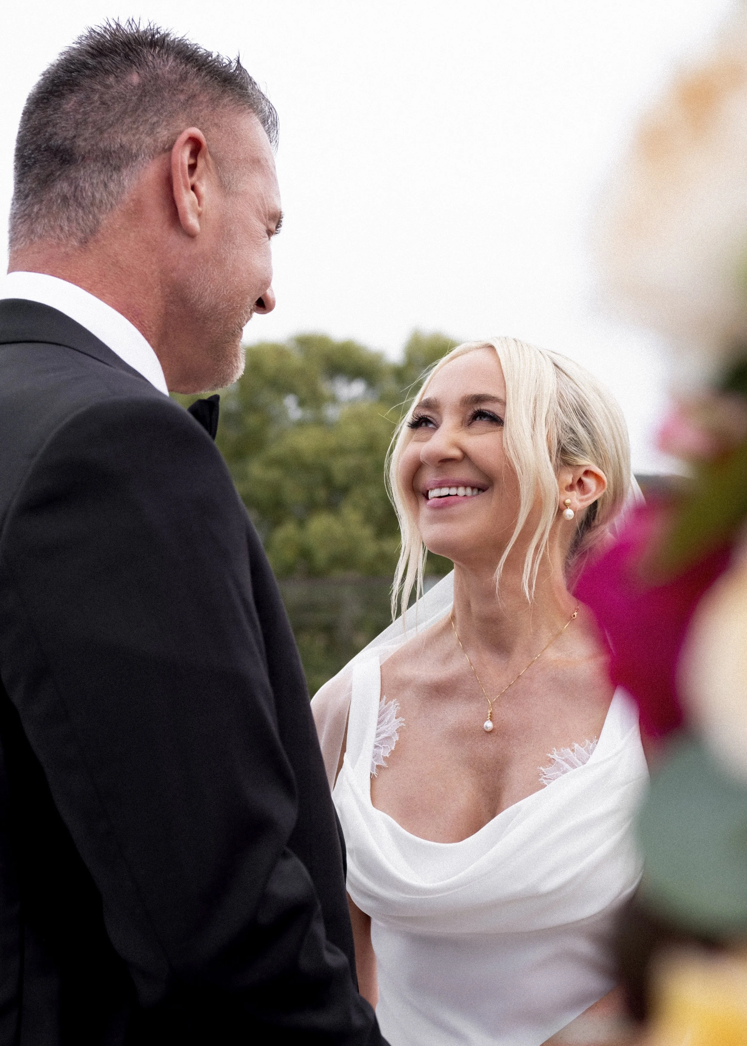 A bride and groom exchange vows outdoors. The bride is smiling and looking at the groom, who is wearing a tuxedo. The background shows a blurred outdoor setting with trees.