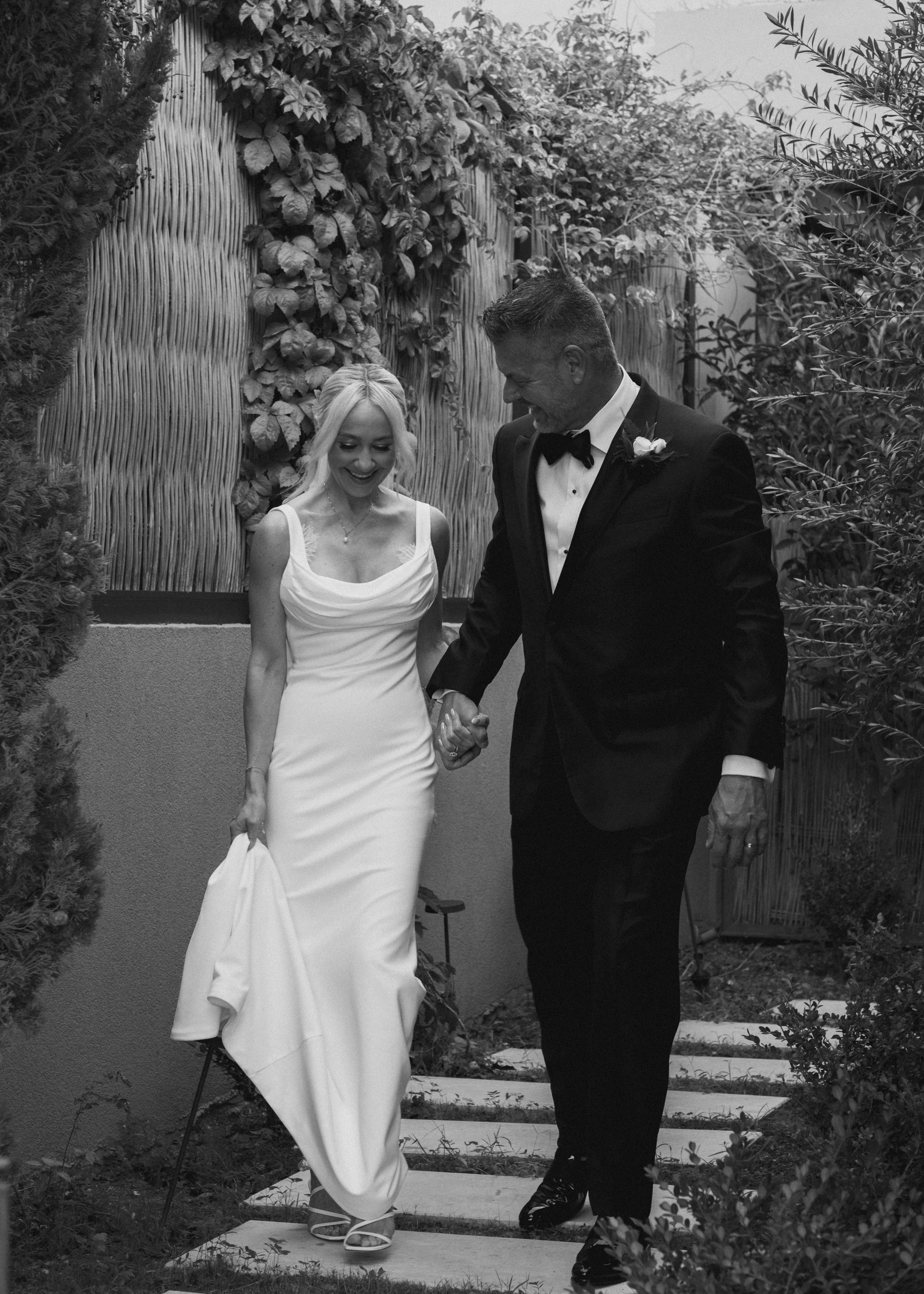 A bride and groom holding hands and walking outdoors, smiling, with the bride in a long white wedding dress and the groom in a black tuxedo.