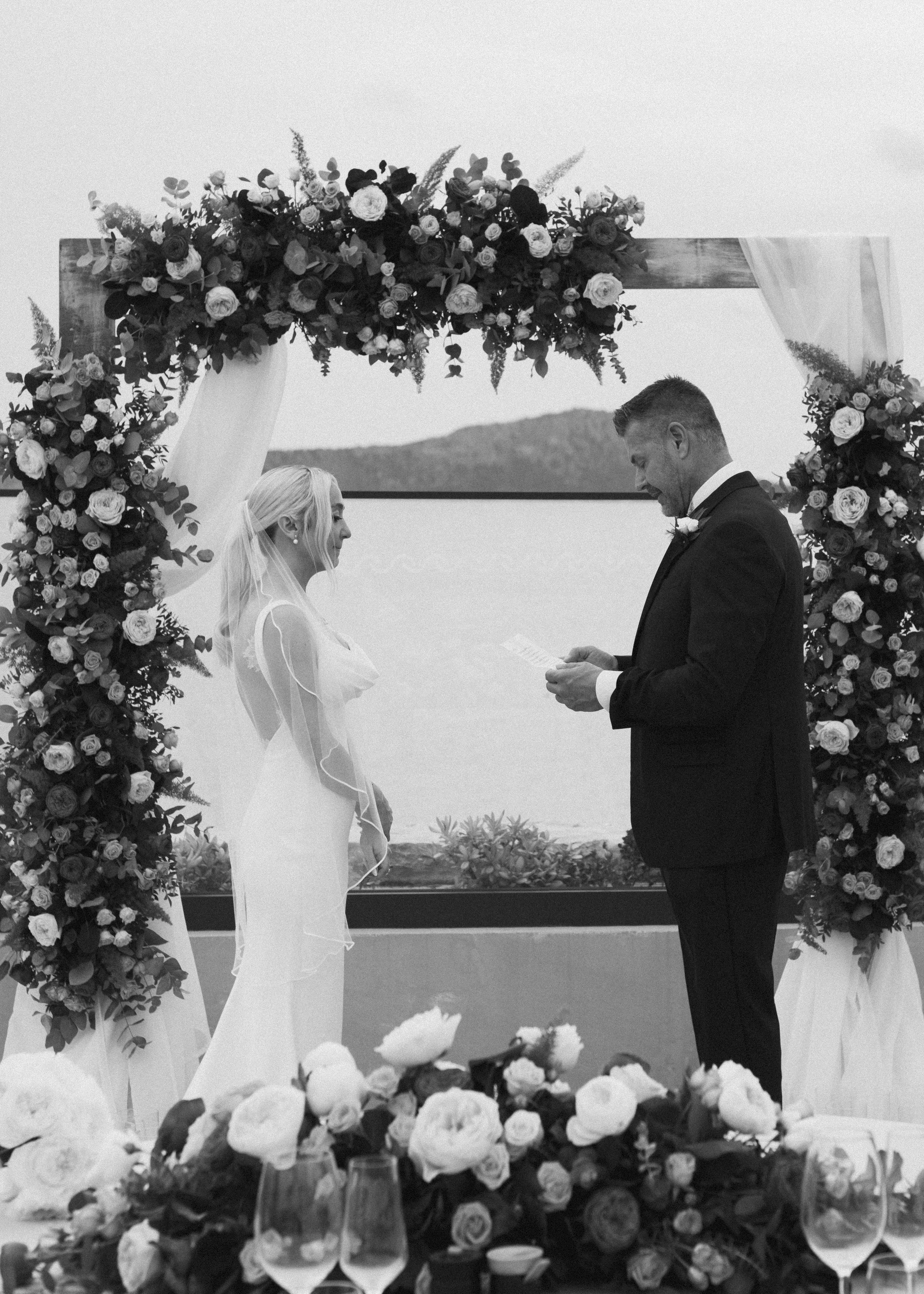 Black and white photo of a wedding ceremony with a bride and groom standing under a floral arch by a body of water, exchanging vows.