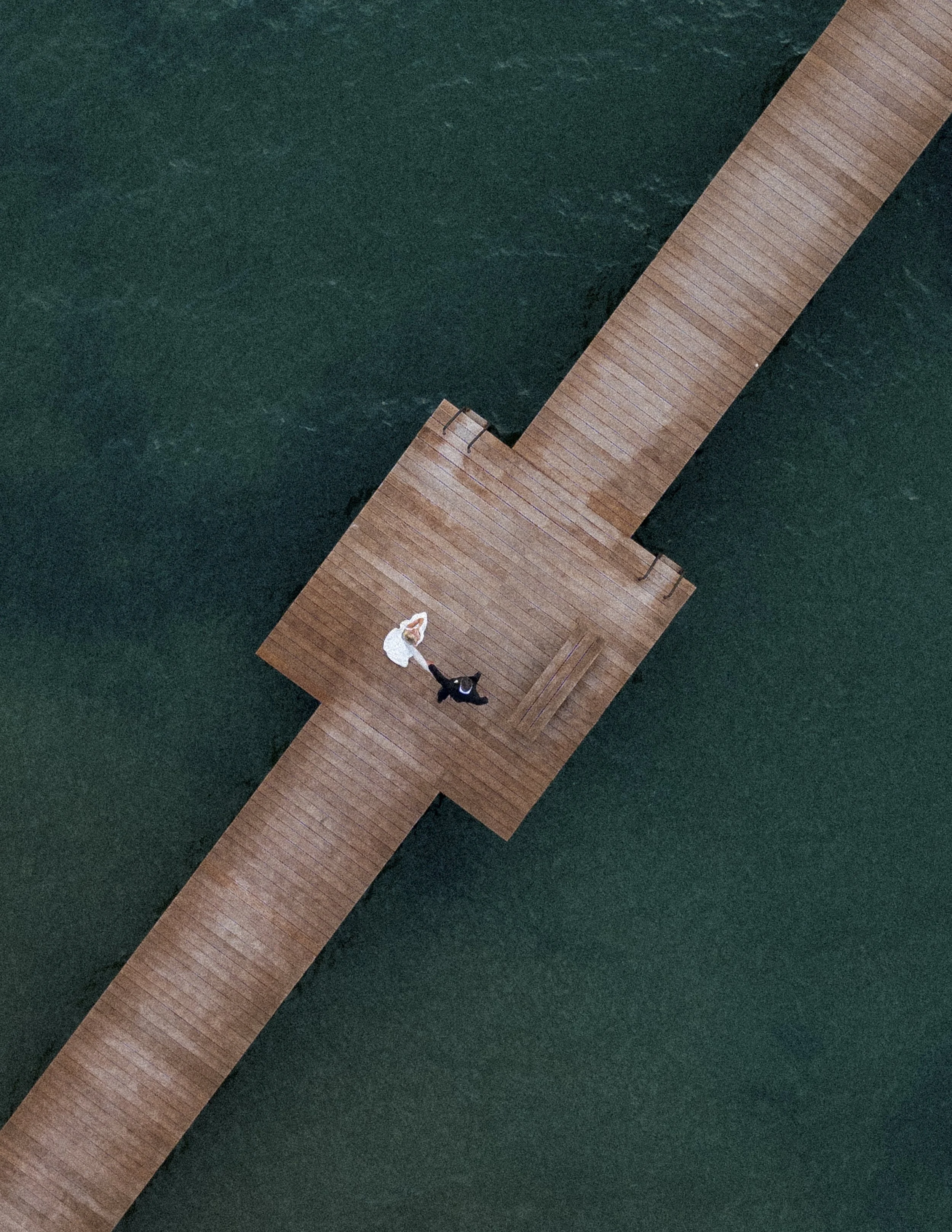 Aerial view of a wooden pier extending into the water with two people sitting at the end, one in white and one in dark clothing.