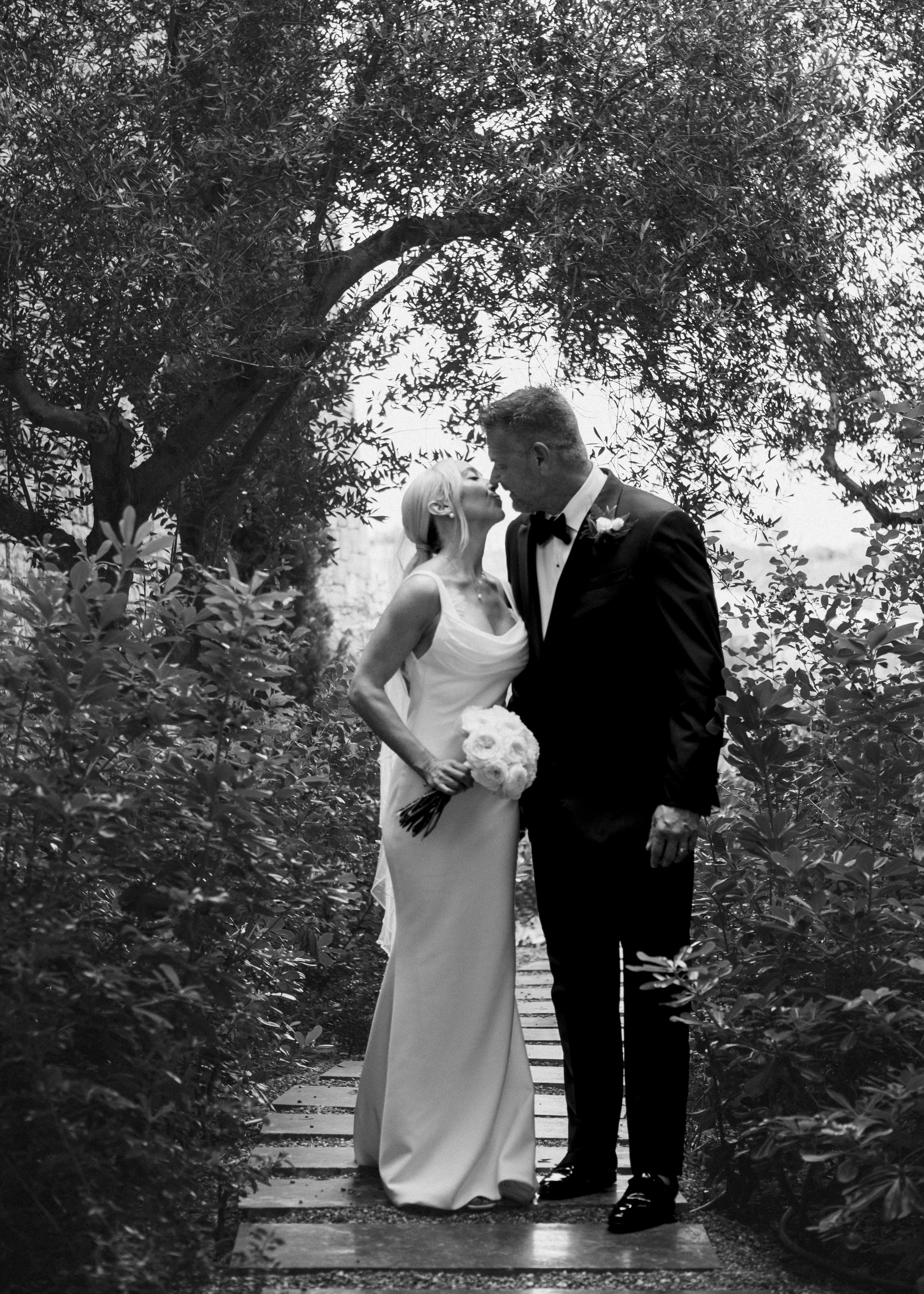 A bride and groom standing on a wooden pathway in a garden, leaning in for a kiss amidst trees and foliage, black and white photograph.
