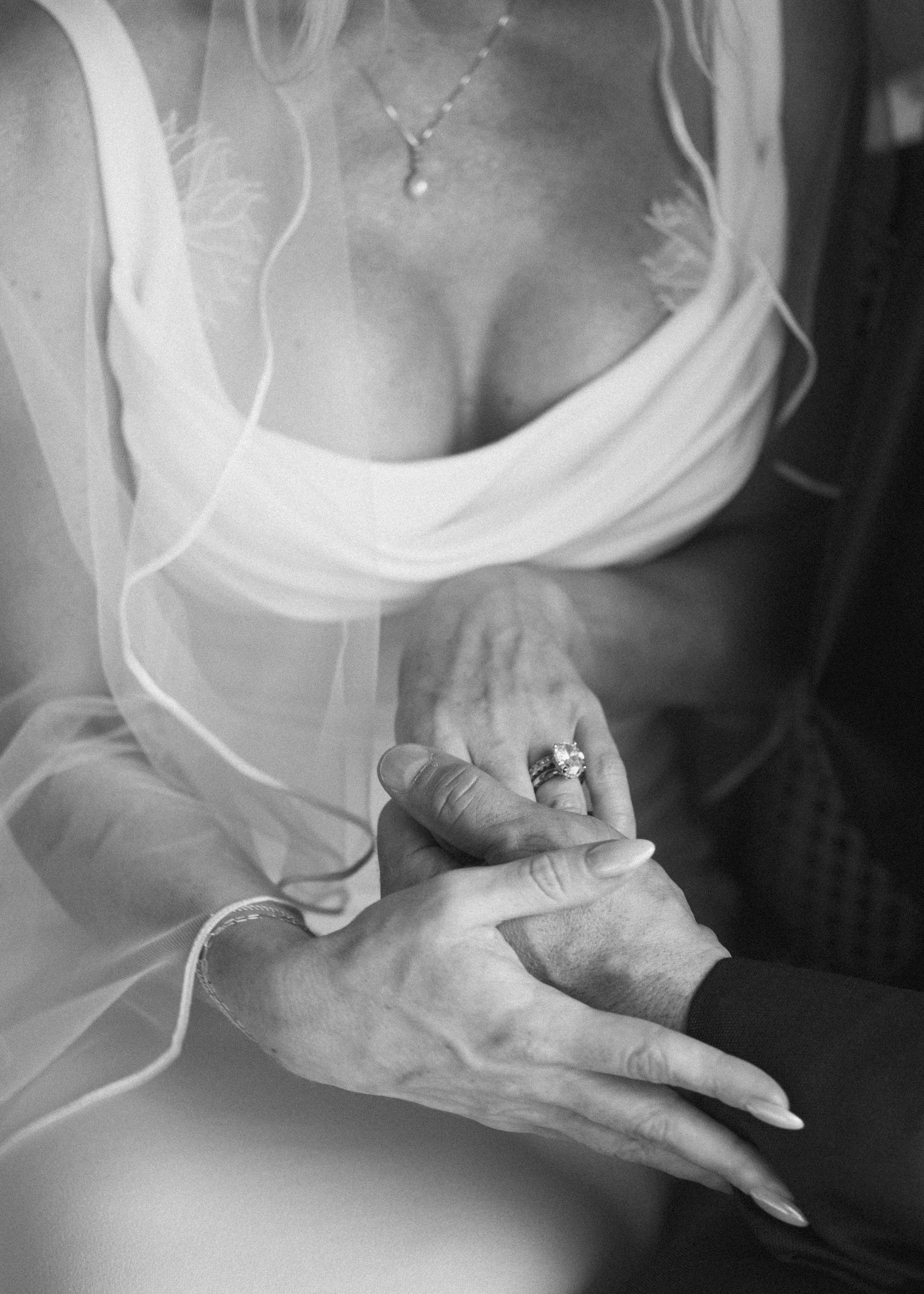A close-up black and white photo of a woman wearing a wedding dress and a necklace, holding hands with an older person, showing an engagement ring on her finger.