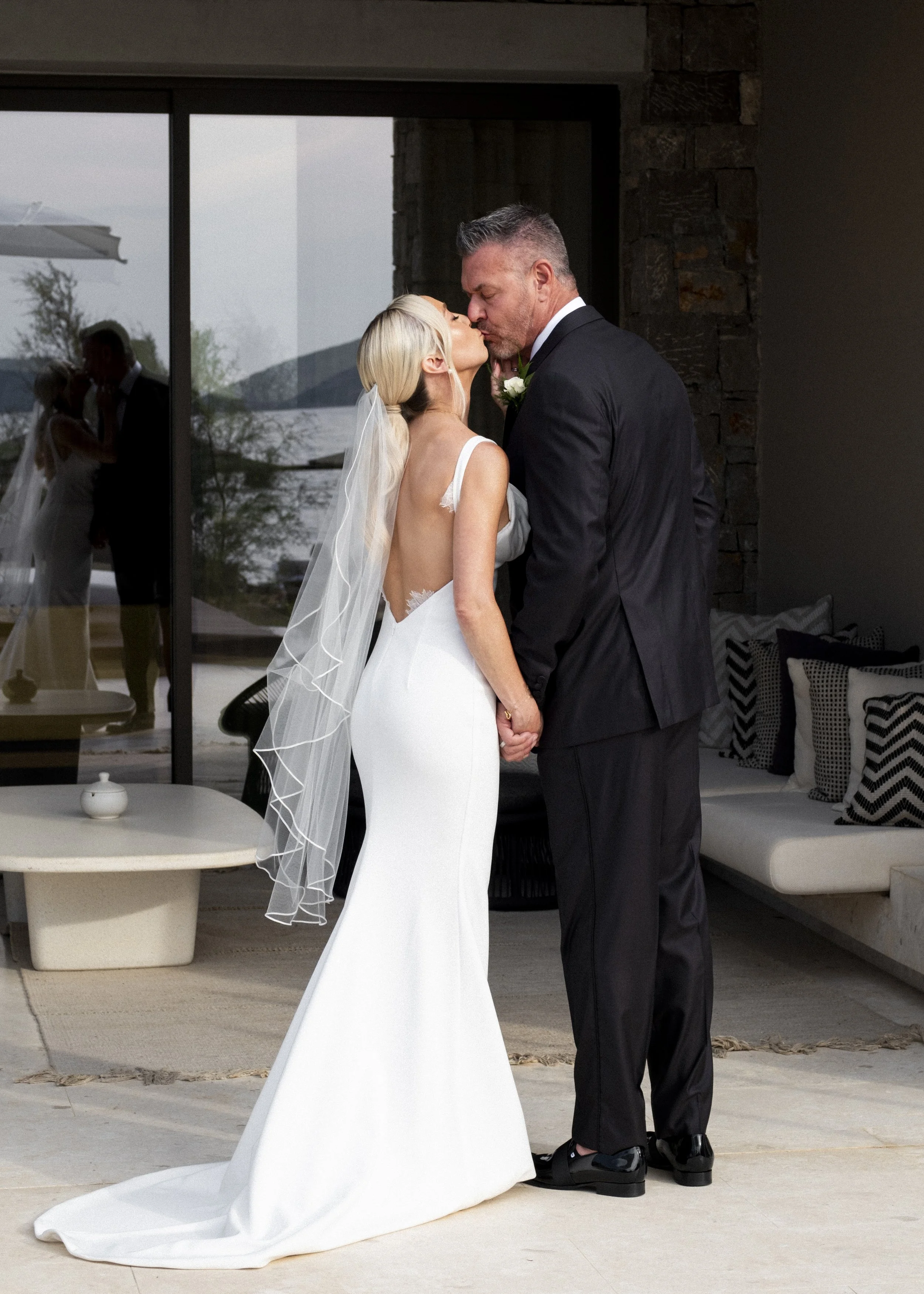 A bride and groom share a kiss during their wedding, holding hands inside a modern venue with large glass windows, a view of a lake, and a stone accent wall.