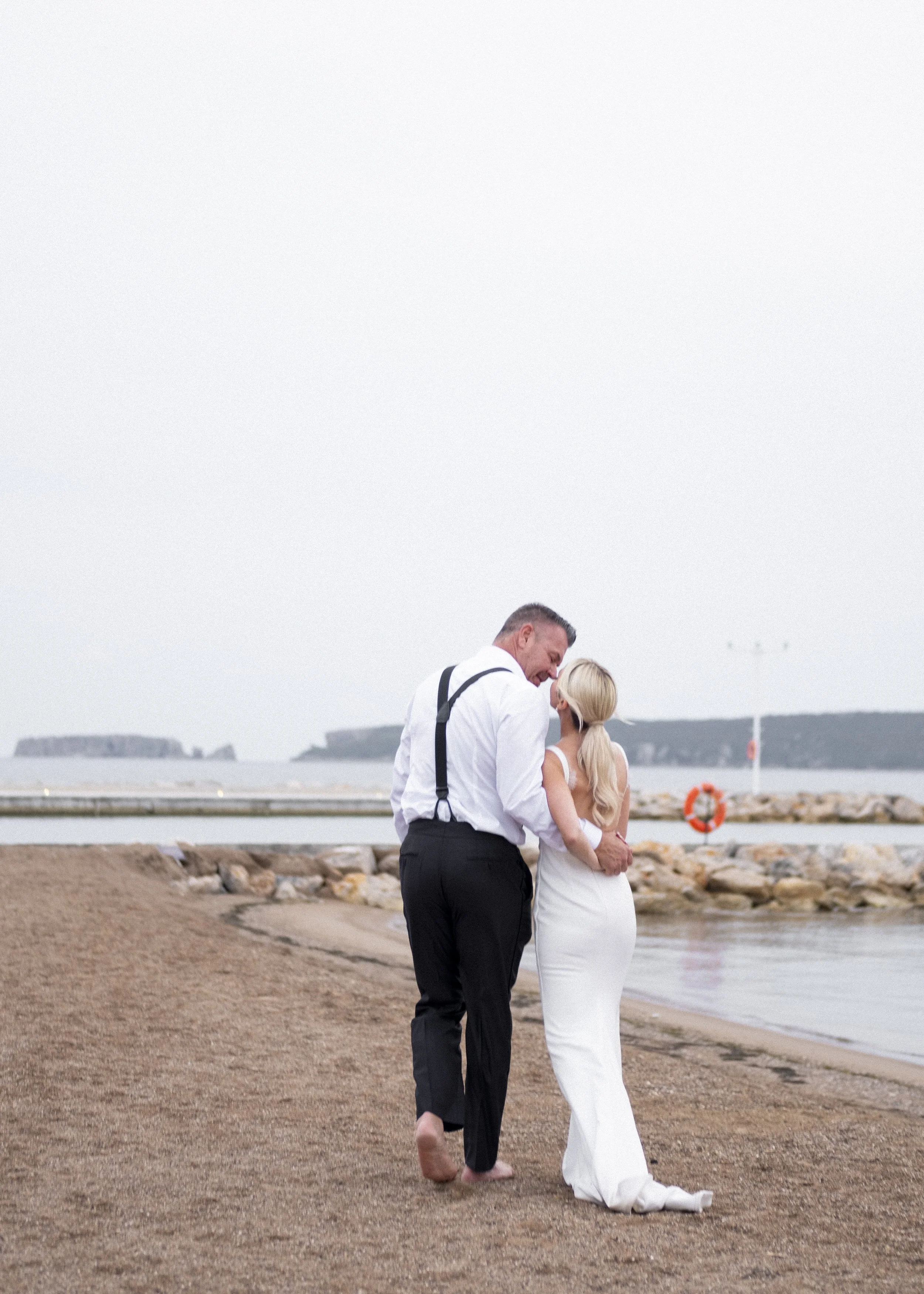A man and woman walking barefoot on the beach, embracing and smiling, with rocky shoreline and water in the background.