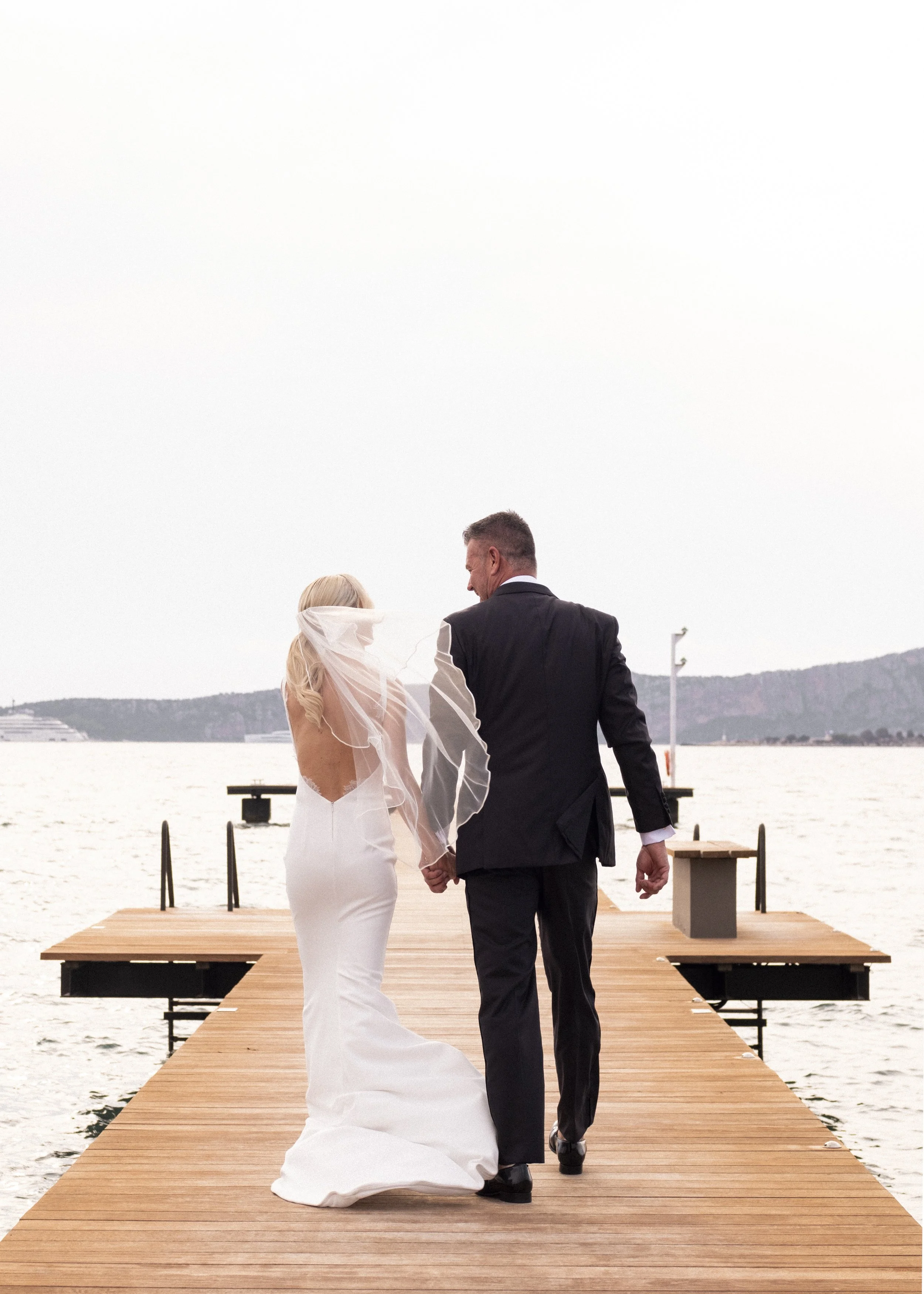 A bride and groom walking hand in hand on a wooden dock by the water, with a scenic mountain view in the background.