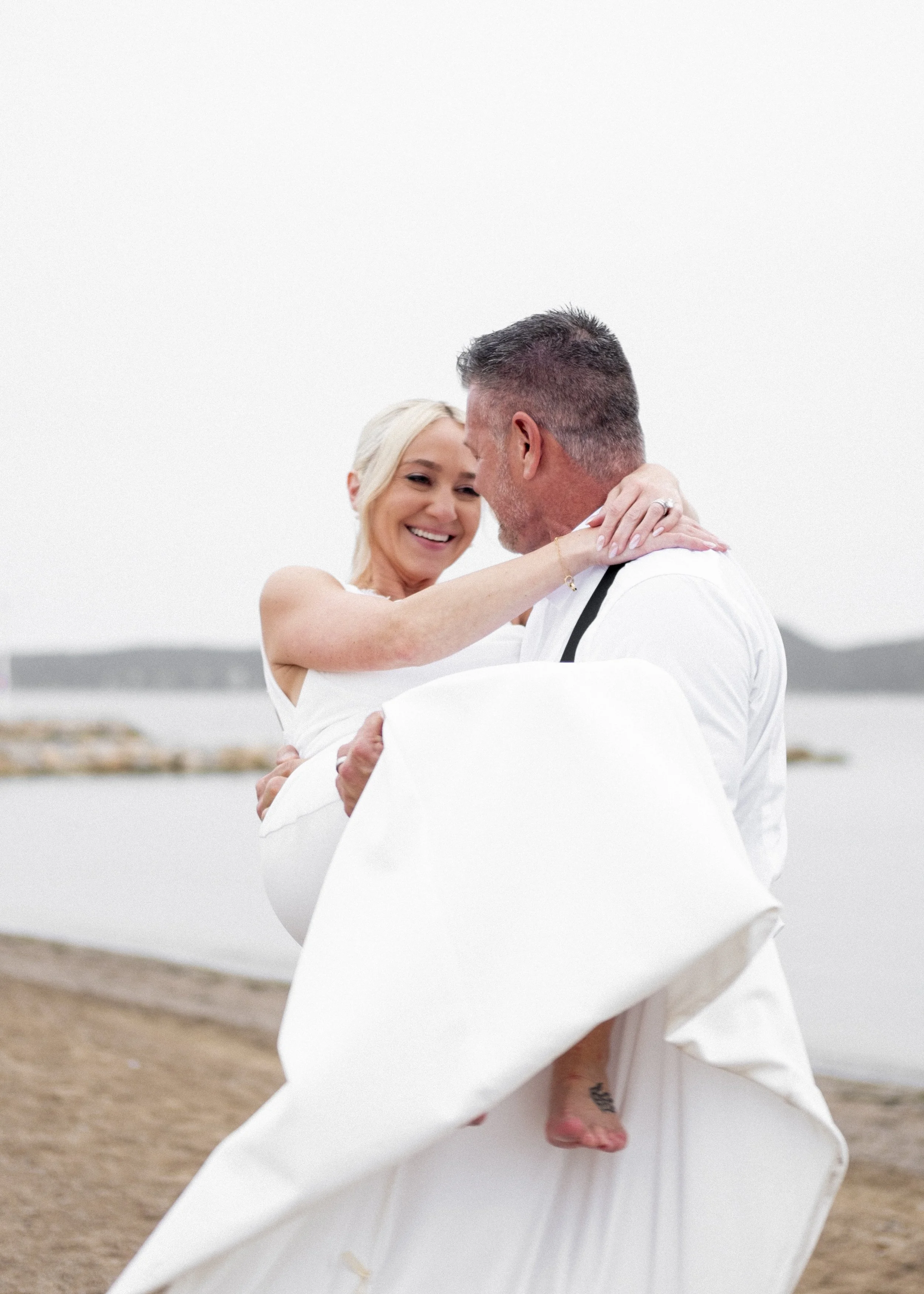 A bride and groom sharing a joyful moment outdoors near a body of water, with the groom holding the bride in his arms.
