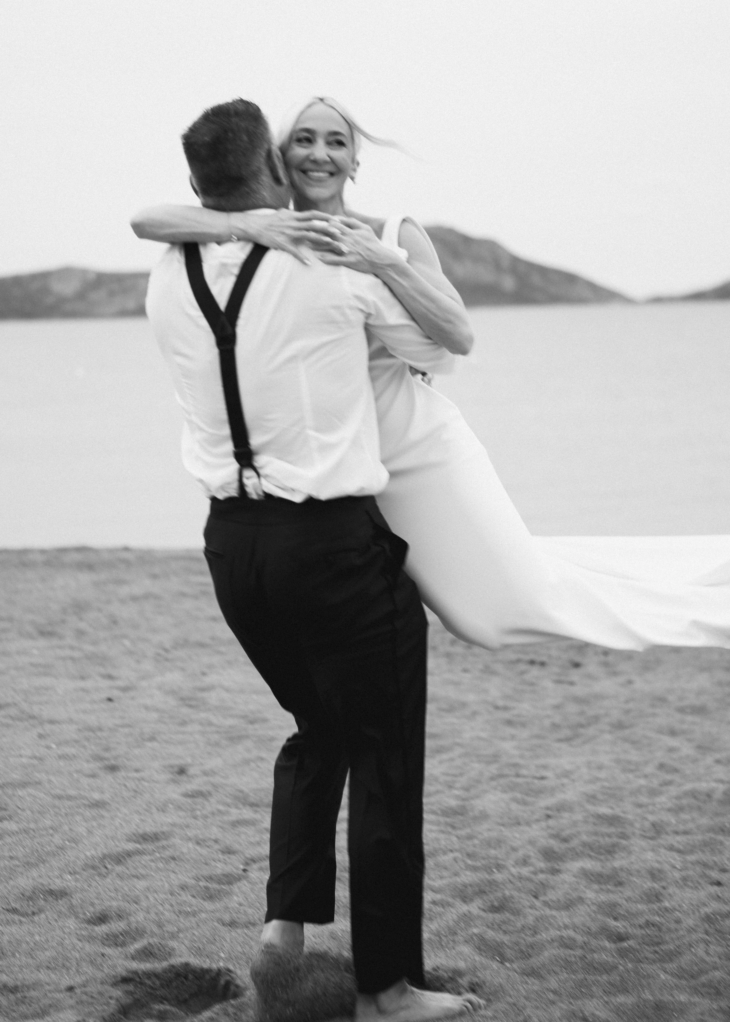 A black and white photo of a smiling woman in a dress being lifted by a man on a beach, with mountains in the background.