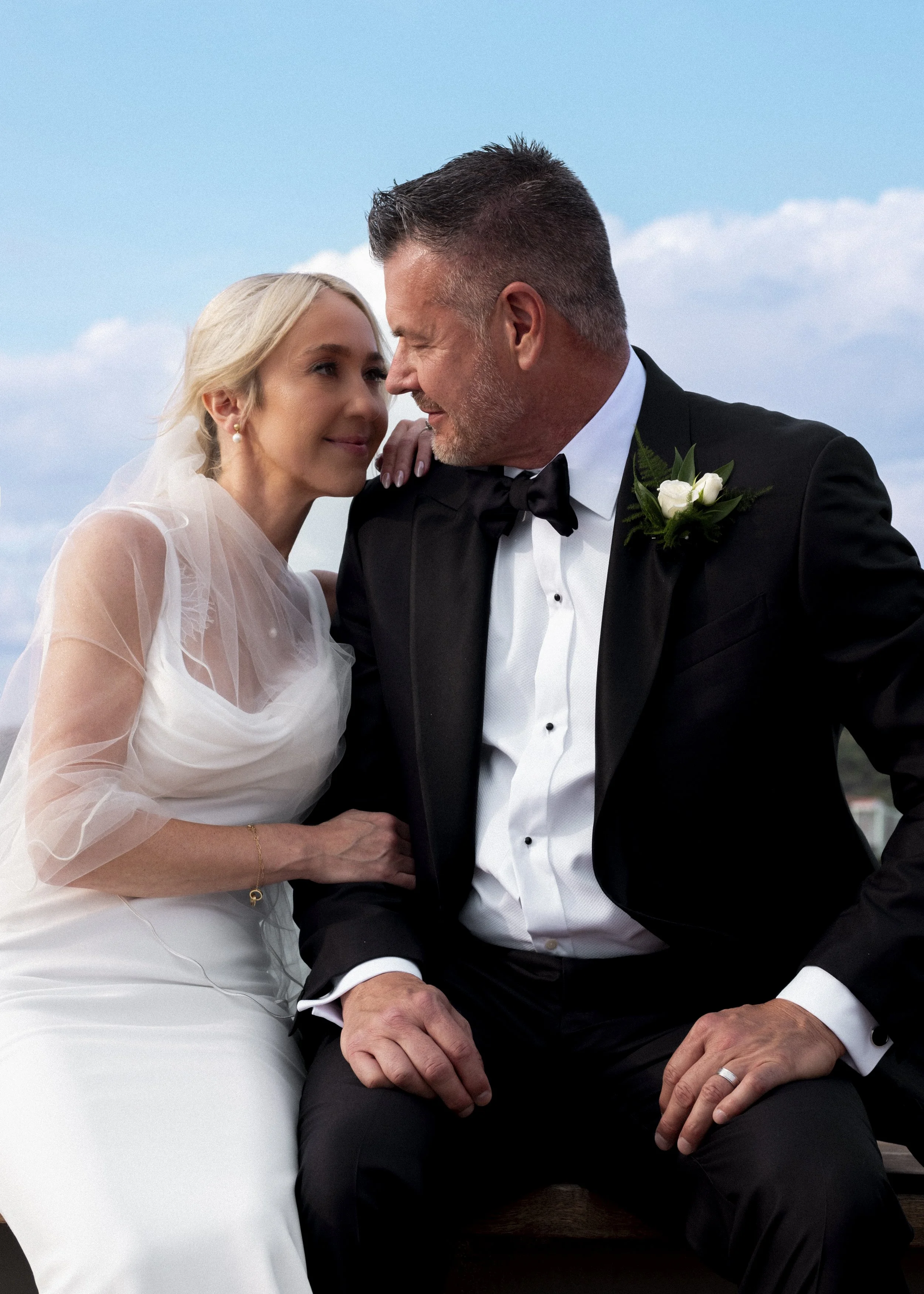 A bride and groom in wedding attire sitting close together outdoors under a partly cloudy sky, sharing an intimate moment.