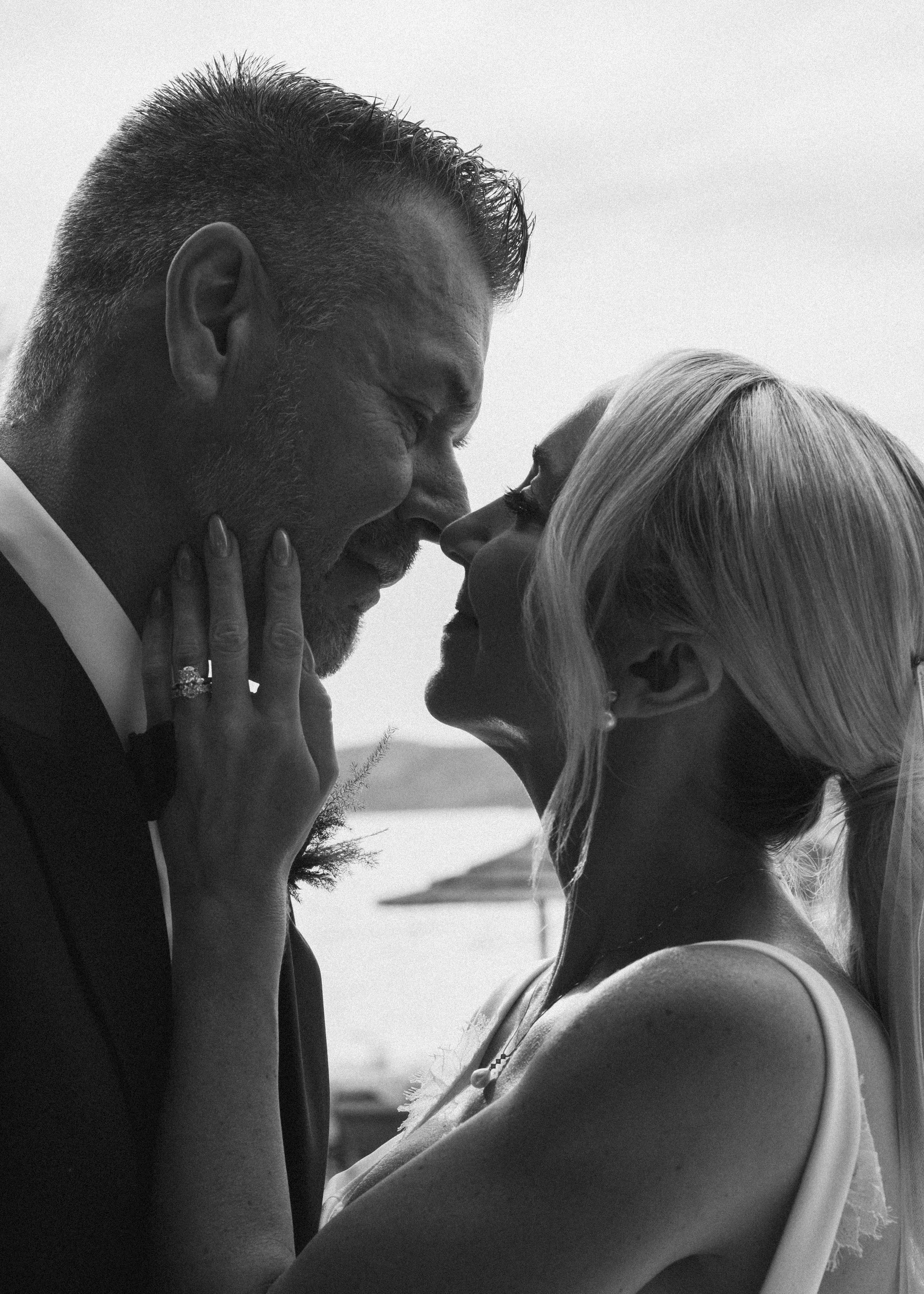 Black and white photo of a man and woman about to kiss, with their faces close and eyes closed, outdoors with water and land in the background.