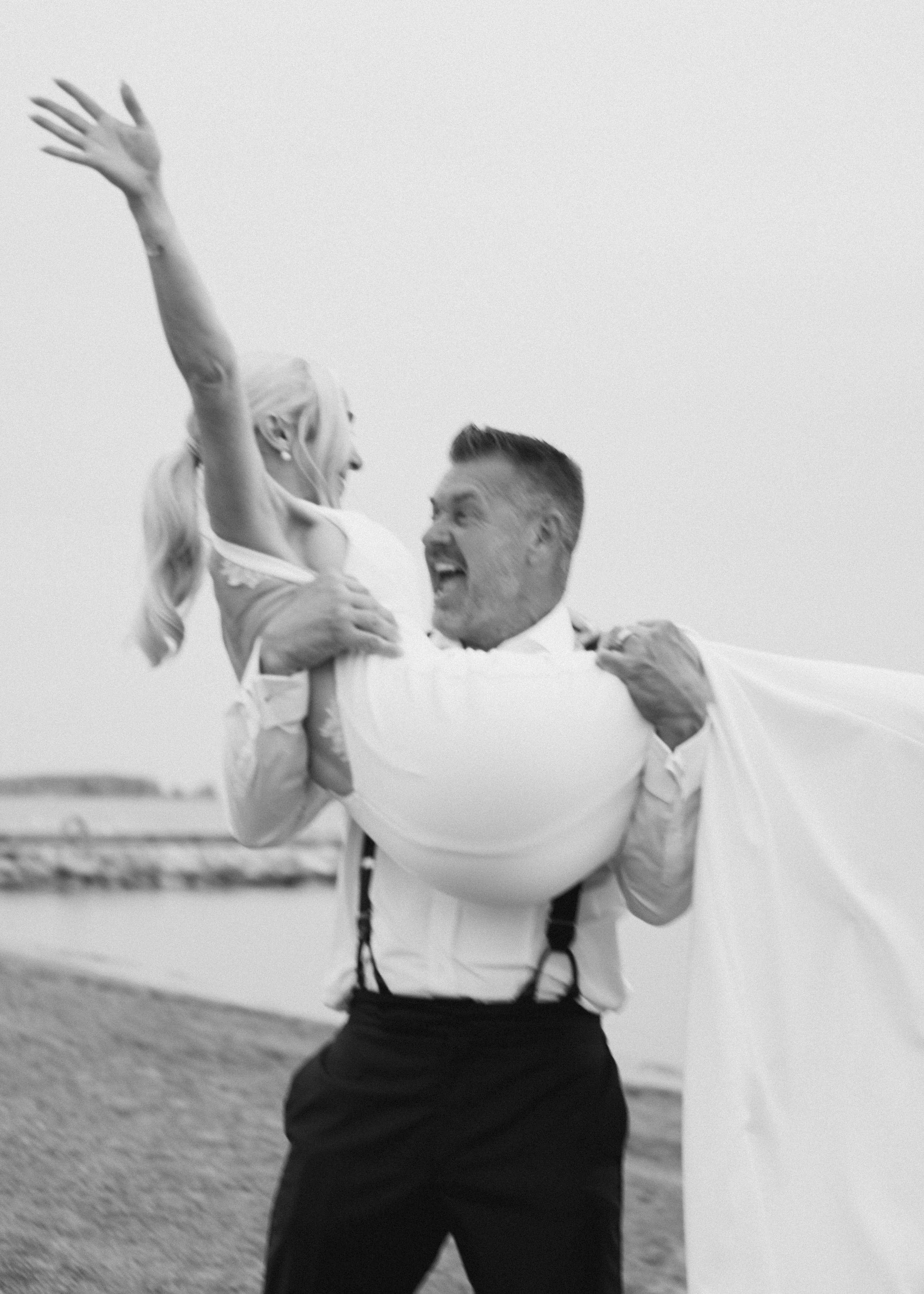 A black and white photo of a man lifting a woman in the air outdoors by a body of water, both smiling and laughing.