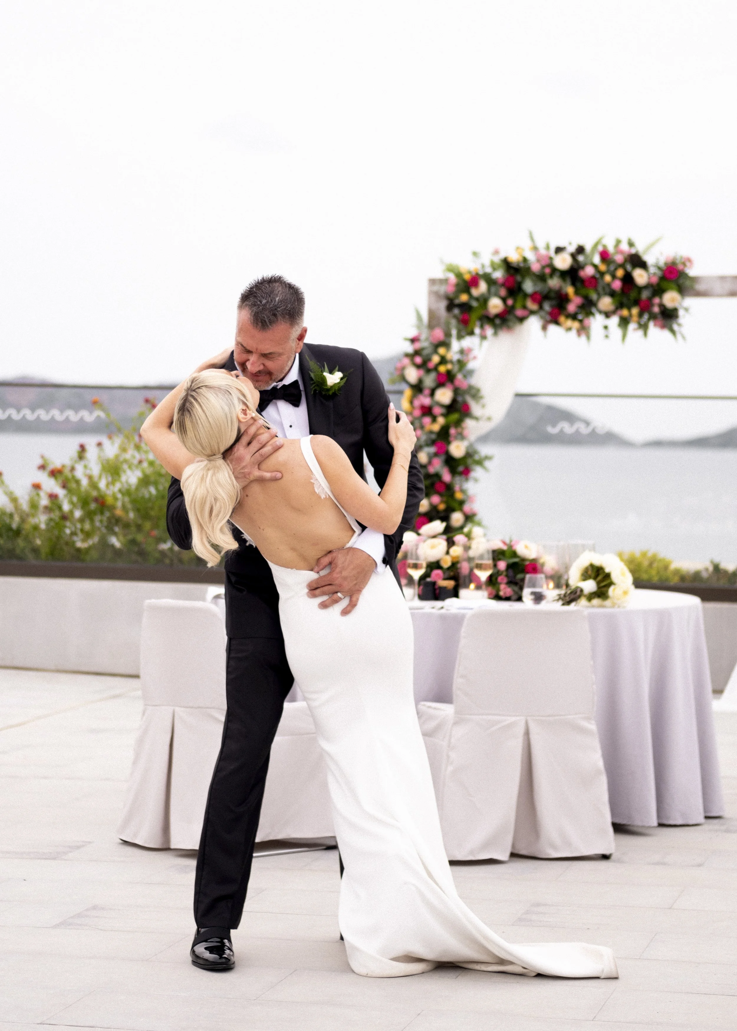 A bride and groom share a dance at their wedding reception outdoors near water, with a decorated floral arch and reception table in the background.