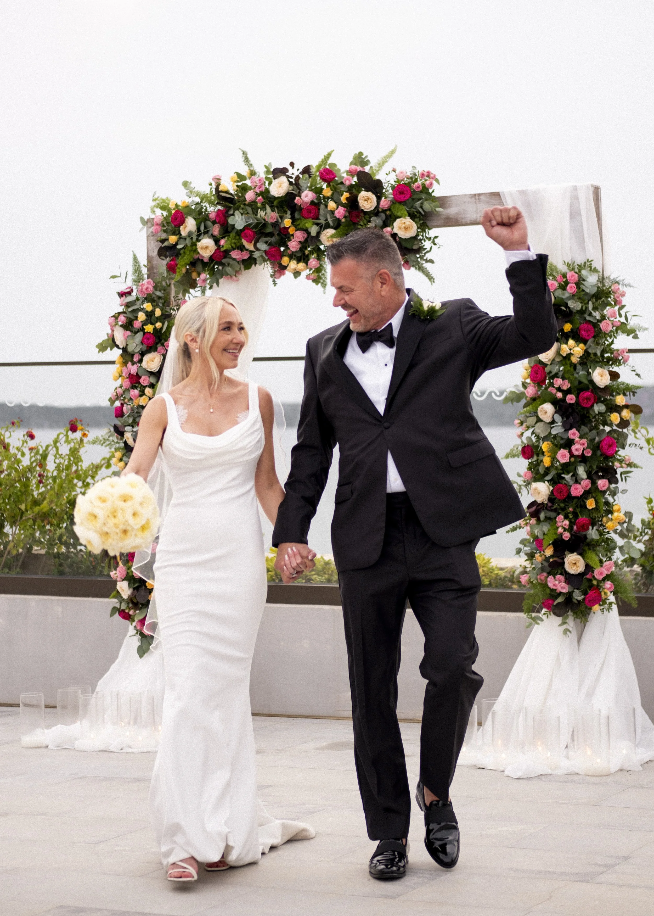 A bride and groom holding hands, celebrating their wedding outdoors under a floral arch, with the groom raising his fist in celebration.