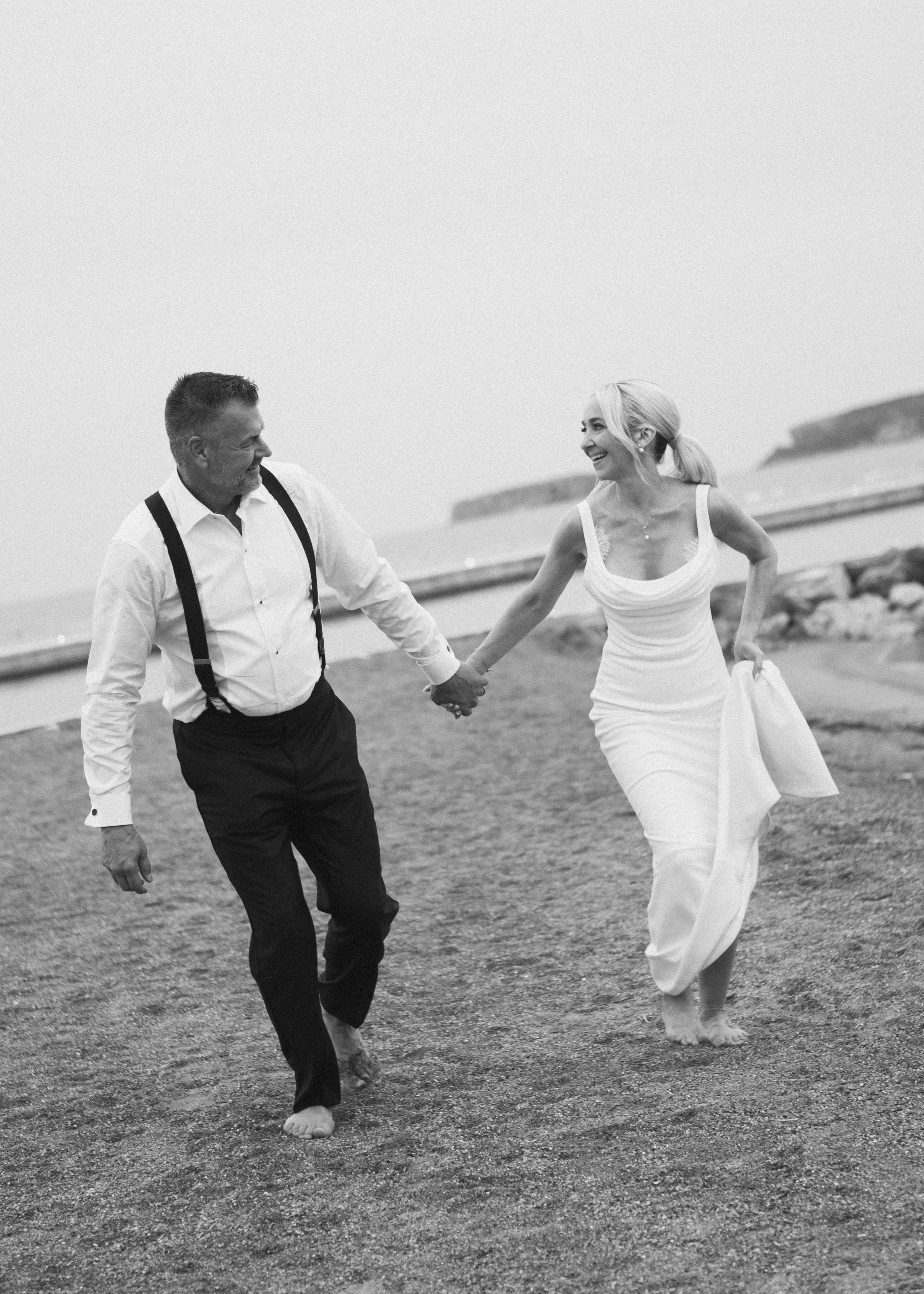 A black and white photo of a man and woman walking barefoot on a beach, holding hands and smiling at each other. The woman is wearing a white dress, and the man is dressed in a white shirt with suspenders and dark trousers.