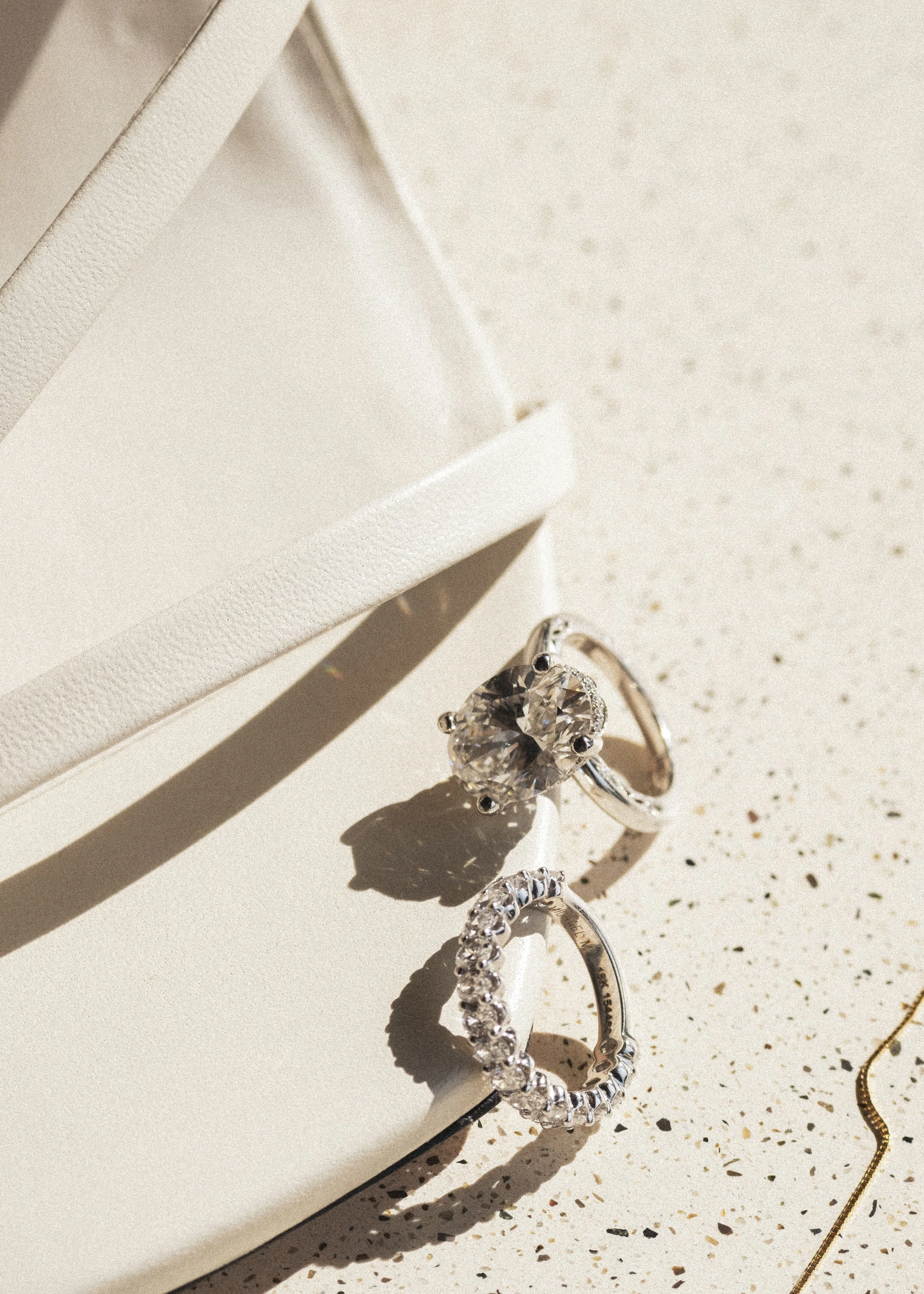 Two elegant rings, one with a large central gemstone and the other a band of smaller stones, are displayed on a white surface next to a white textured box or jewelry holder.