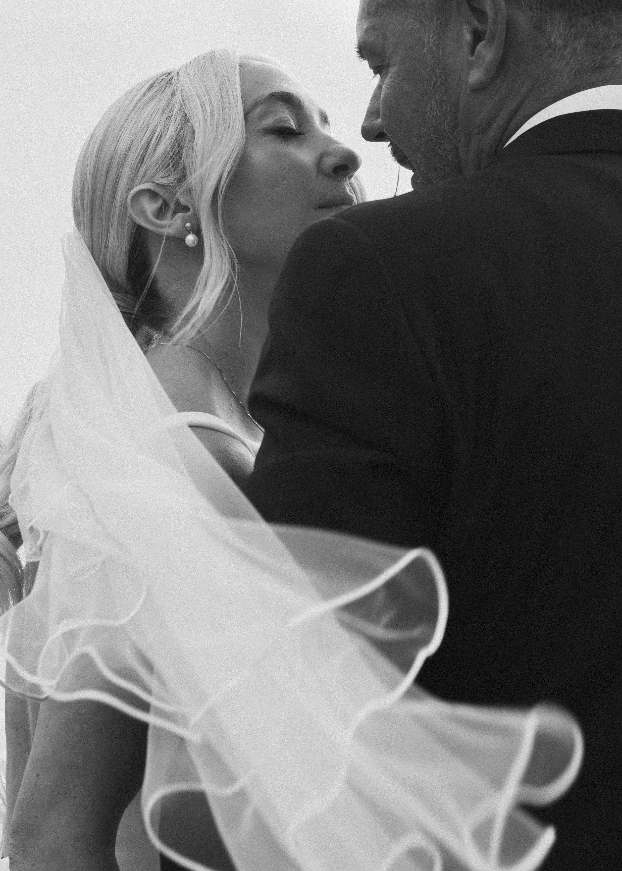 A black and white photo of a couple in a close, intimate moment, possibly during a wedding, with the woman's eyes closed and the man leaning in towards her.