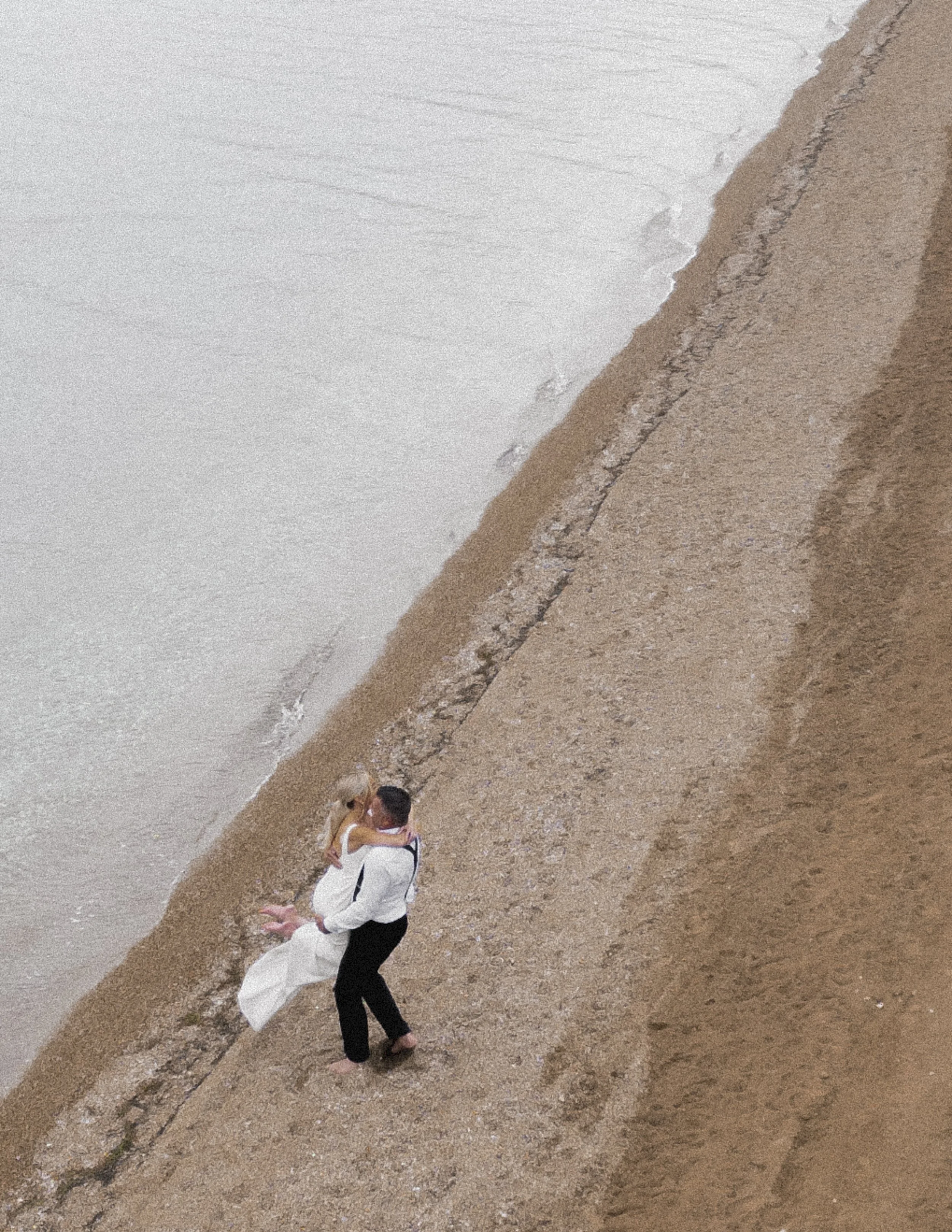 A couple in wedding attire embracing on a sandy beach near the shoreline, seen from above.