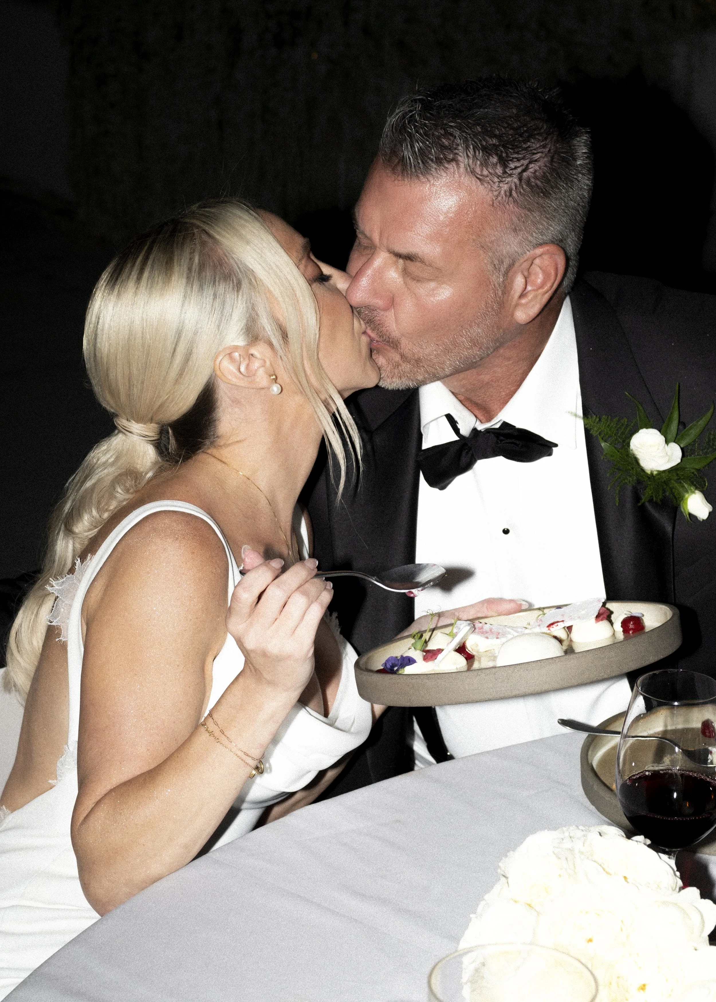 A man in a tuxedo and a woman in a white dress kiss while celebrating at a wedding reception. The woman is holding a spoon and a plate with dessert, and there are glasses of red wine and a floral arrangement on the table.
