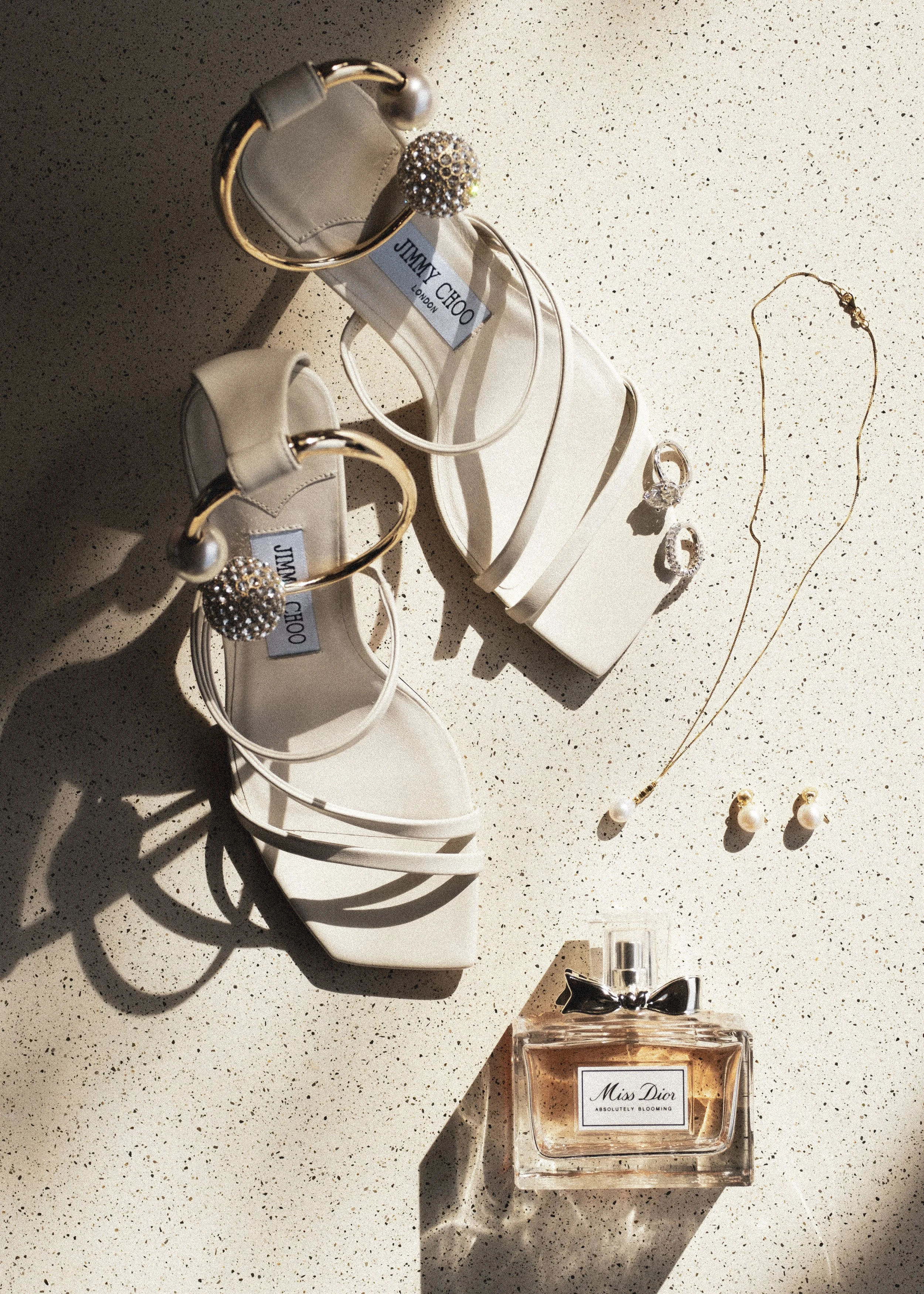 White high-heeled sandals with embellished ankle straps, a gold necklace with pearls, matching pearl earrings, a silver ring, and a bottle of Miss Dior perfume arranged on a speckled surface.