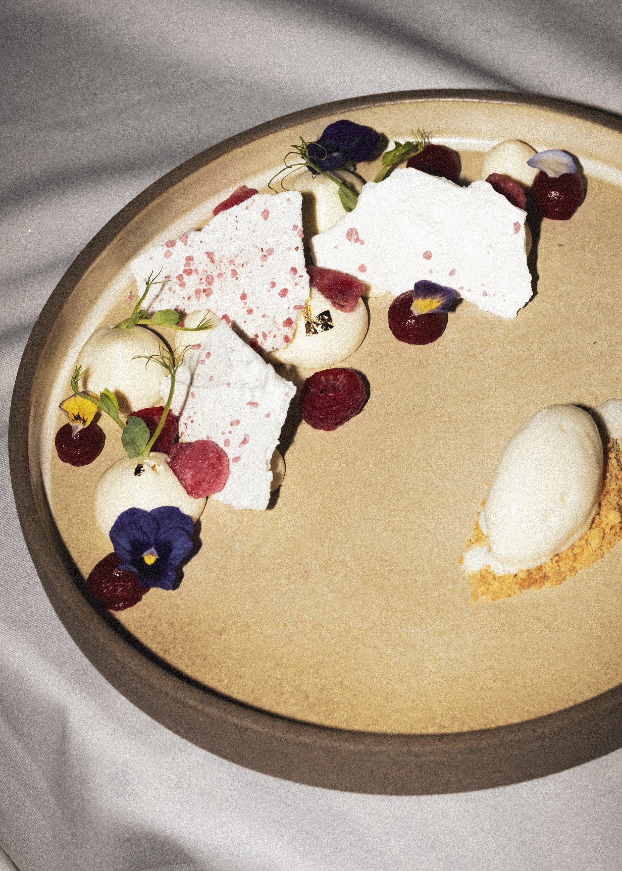 A plated dessert with white chocolate shards, raspberries, cherries, edible flowers, and ice cream on a beige plate.