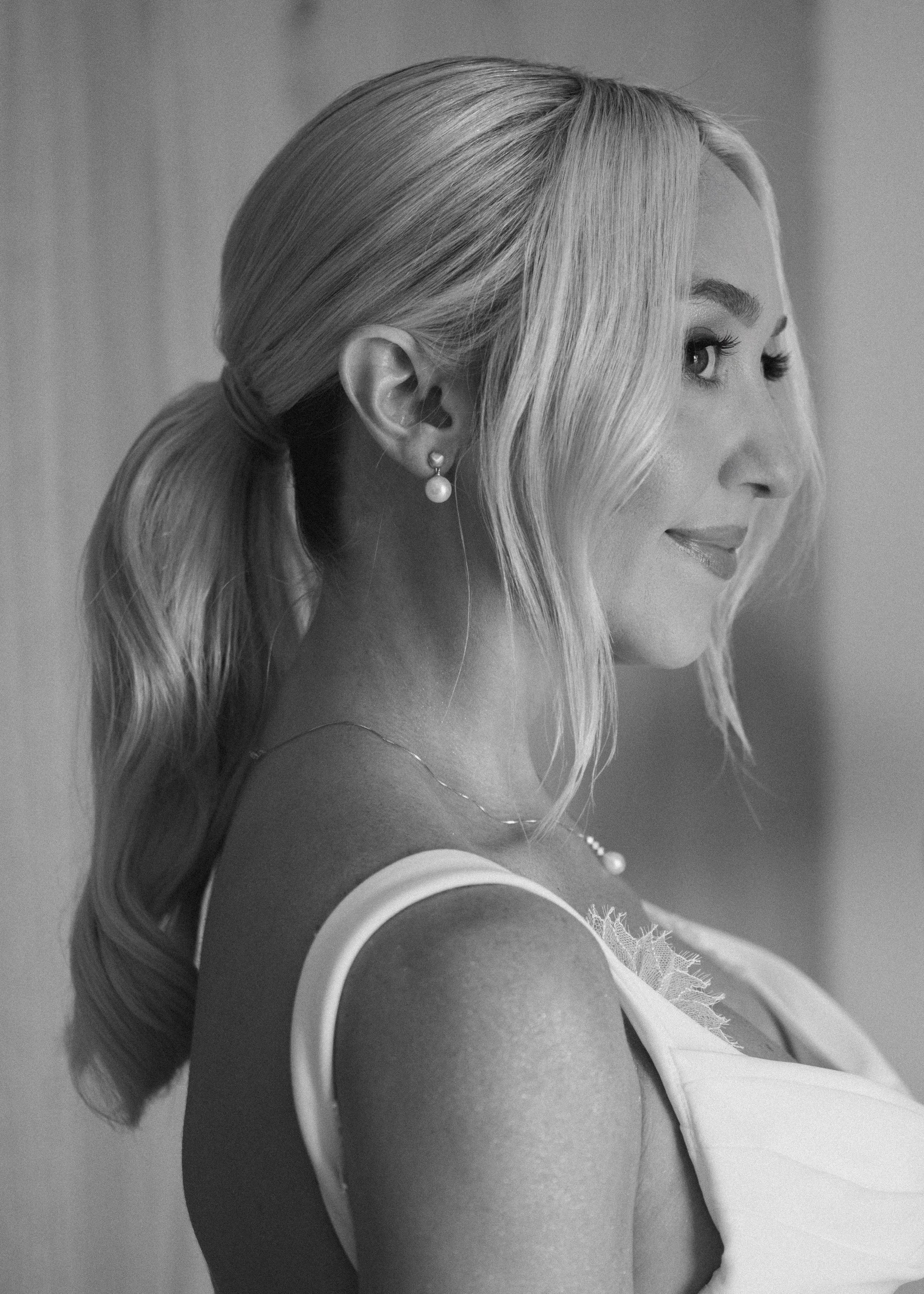 Black and white photograph of a woman with light-colored hair styled in loose waves, wearing pearl earrings and a necklace, looking to her right with a slight smile, while dressed in a sleeveless top.