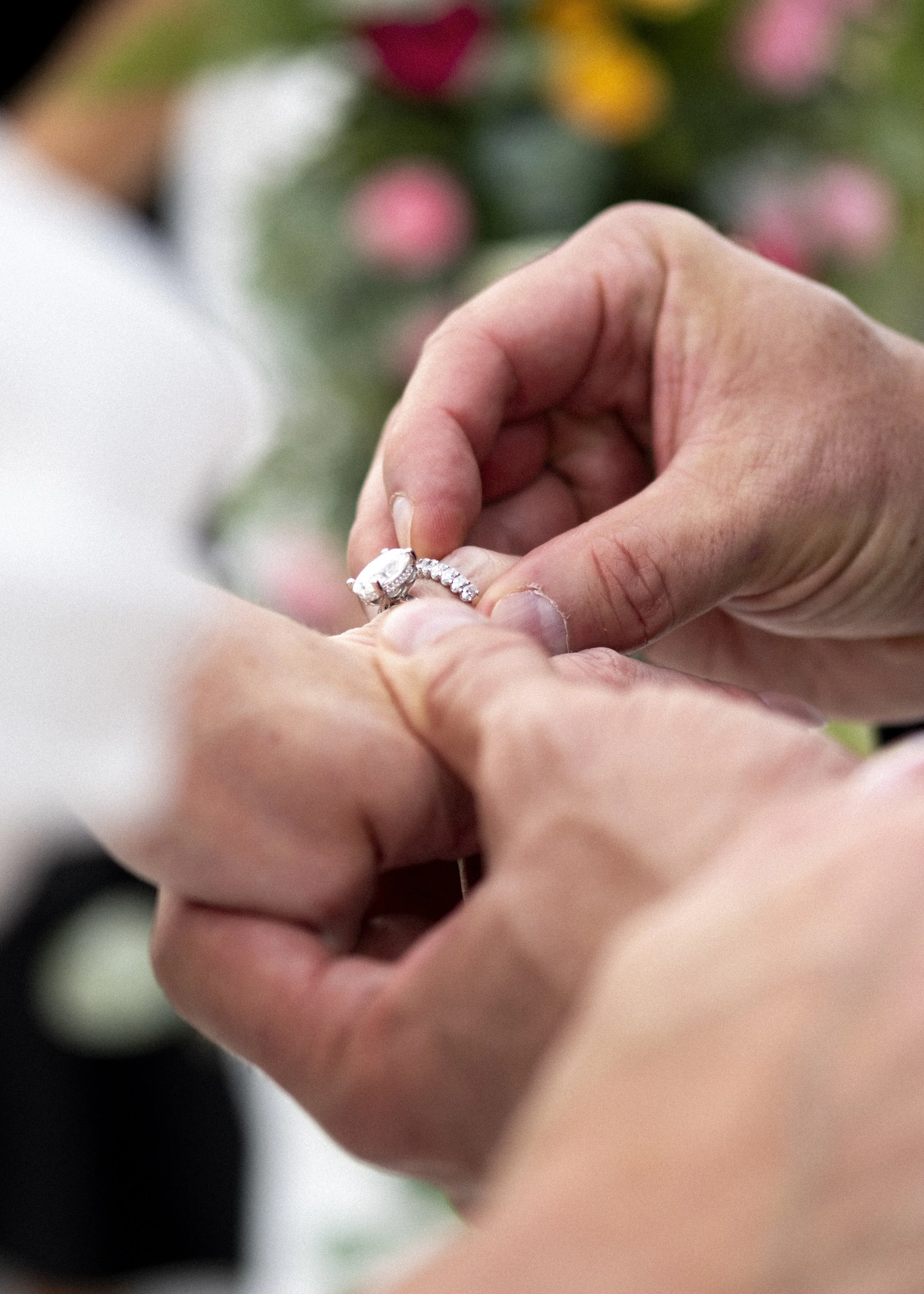 A person is placing a diamond engagement ring onto another person's finger during a wedding or engagement ceremony, with a blurred bouquet of pink flowers in the background.