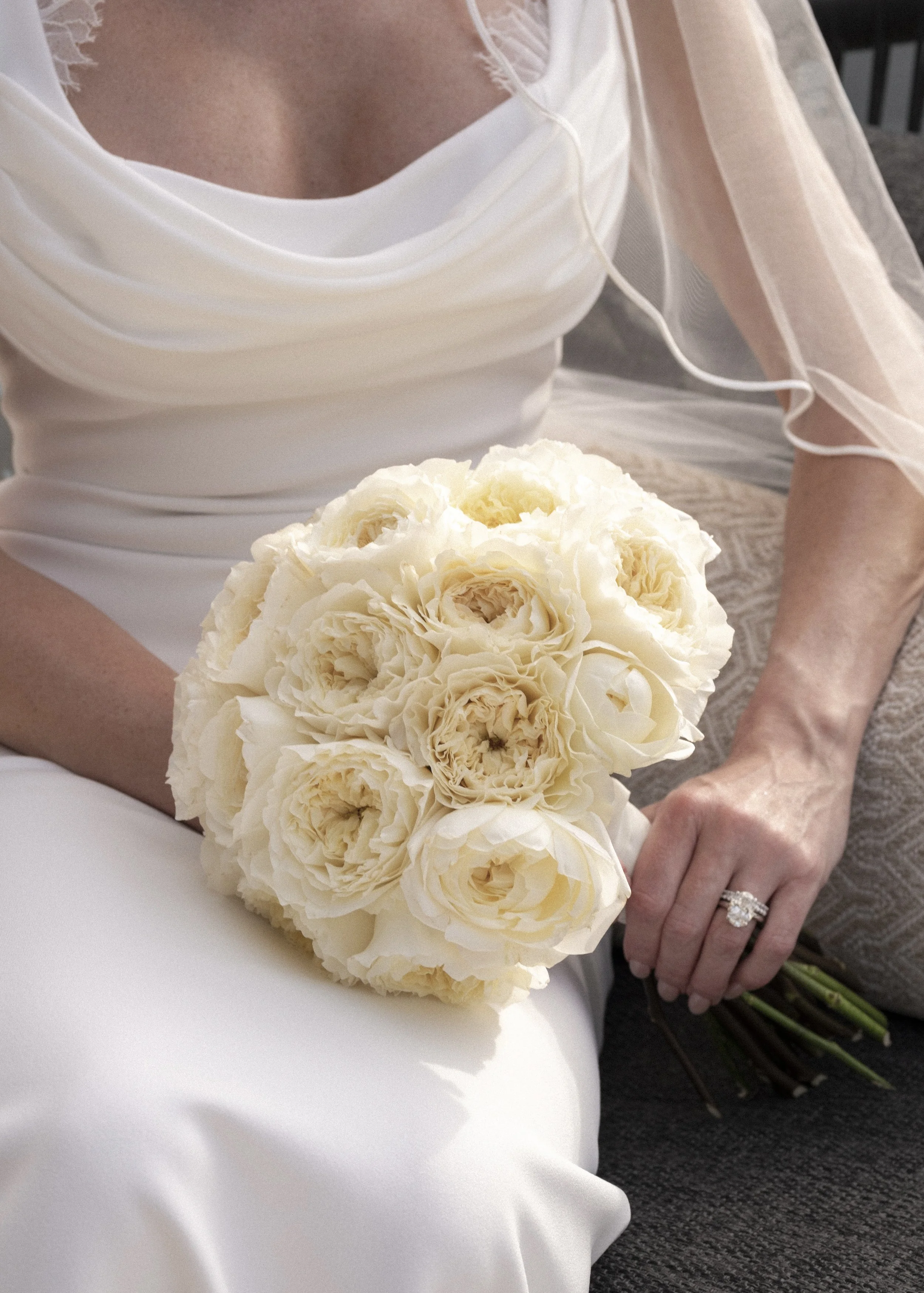 A bride in a white dress holding a bouquet of cream-colored roses, sitting on a dark gray surface with her left hand resting on her lap, showing a diamond engagement ring. Her face is not visible.