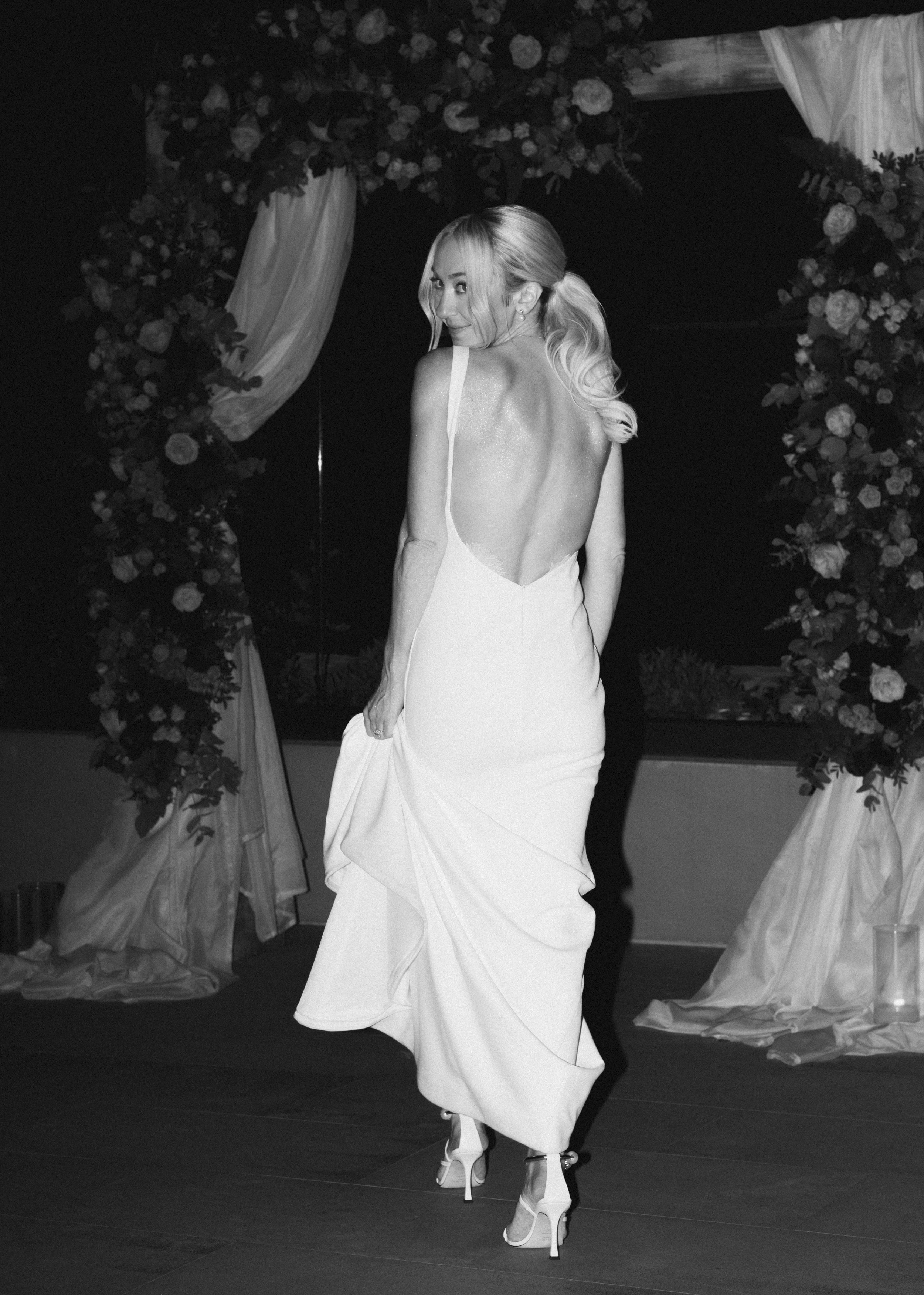 A woman in a backless gown and high heels poses at a decorated event, with floral arch and drapes in the background.