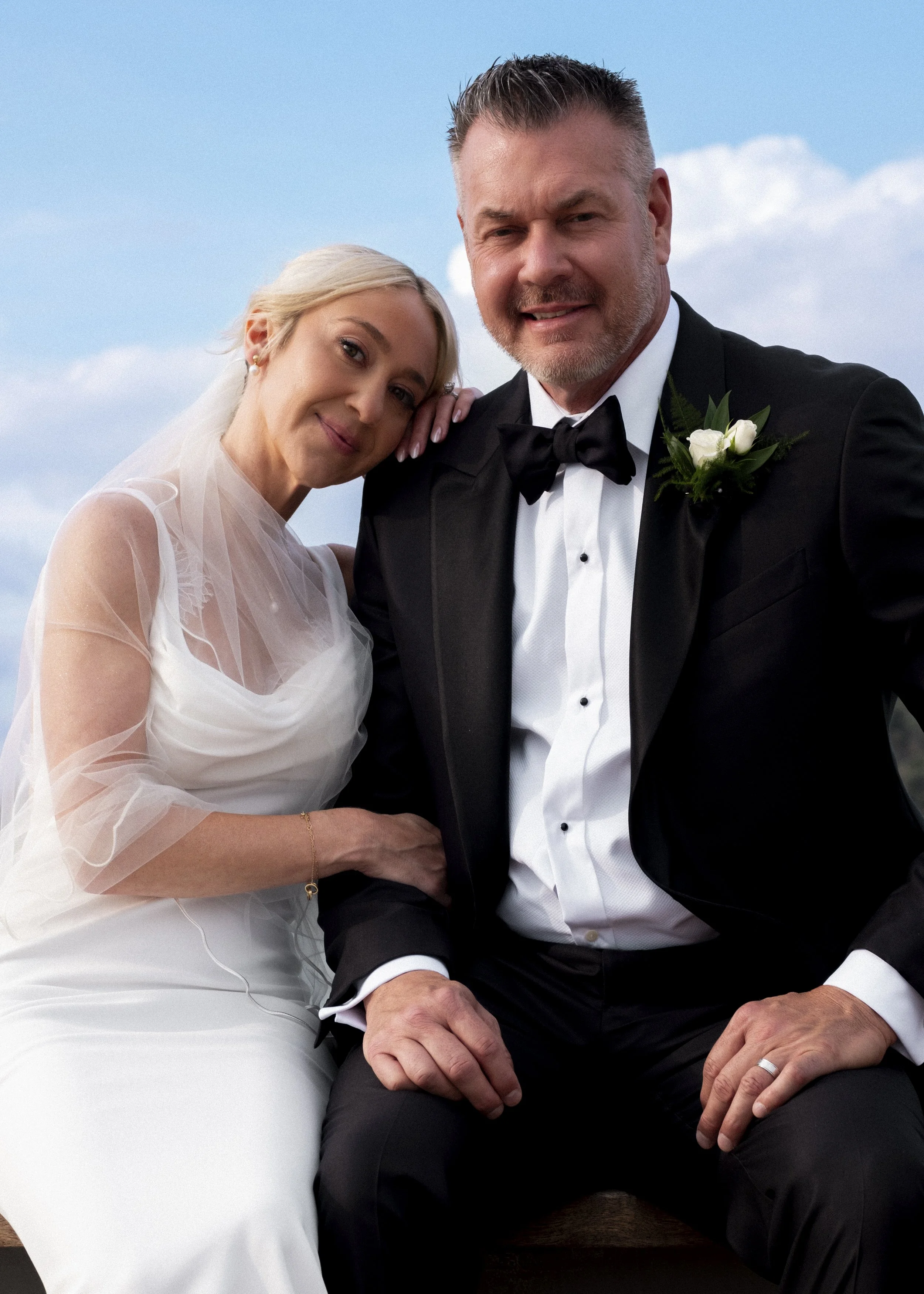 A bride and groom sitting close outdoors, smiling at the camera. The bride is in a white wedding gown with sheer sleeves, and the groom is in a black tuxedo with a bow tie and boutonniere, against a blue sky with clouds.