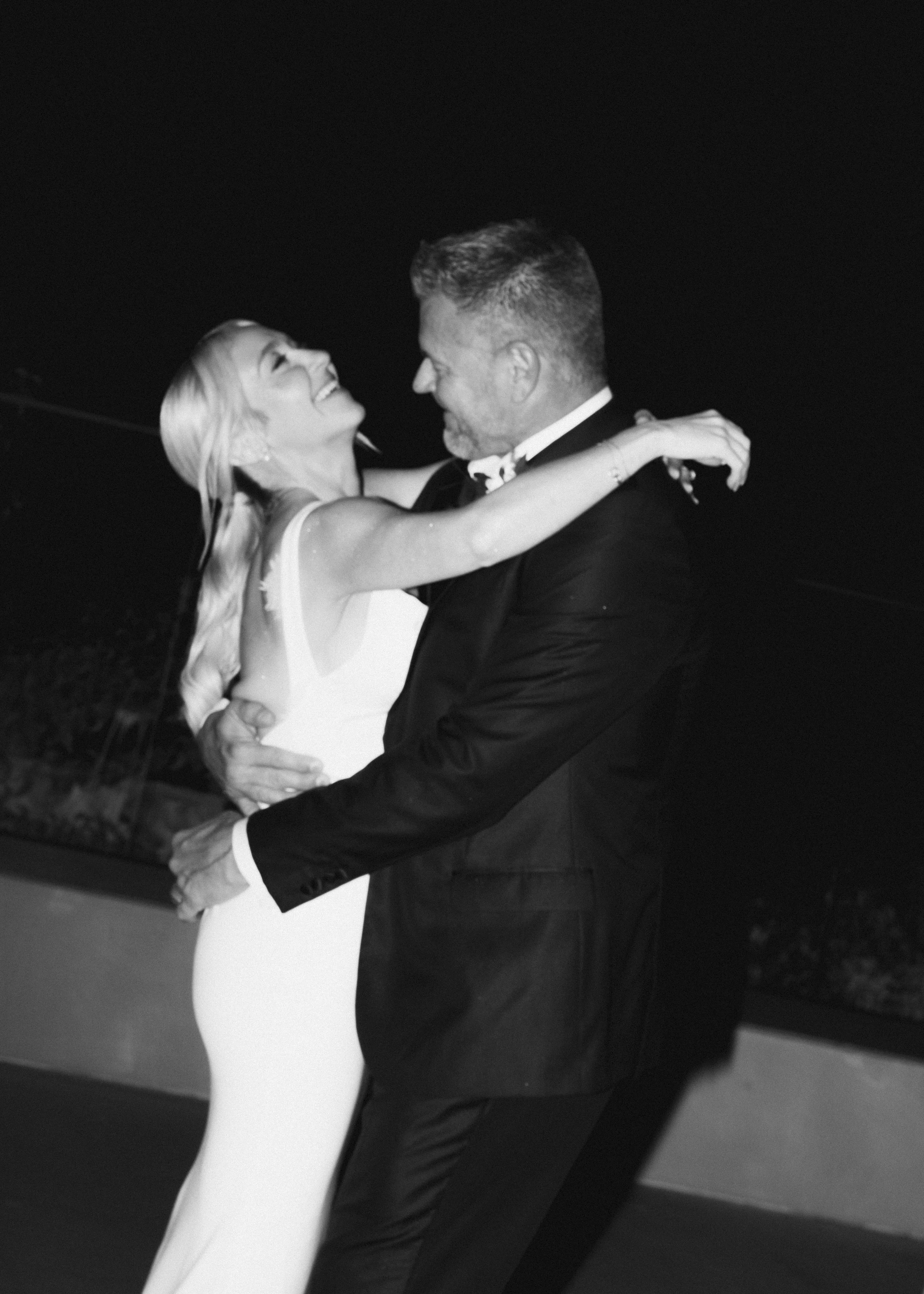 A black-and-white photo of a smiling woman in a sleeveless dress dancing closely with a man in a suit, outdoors at night.