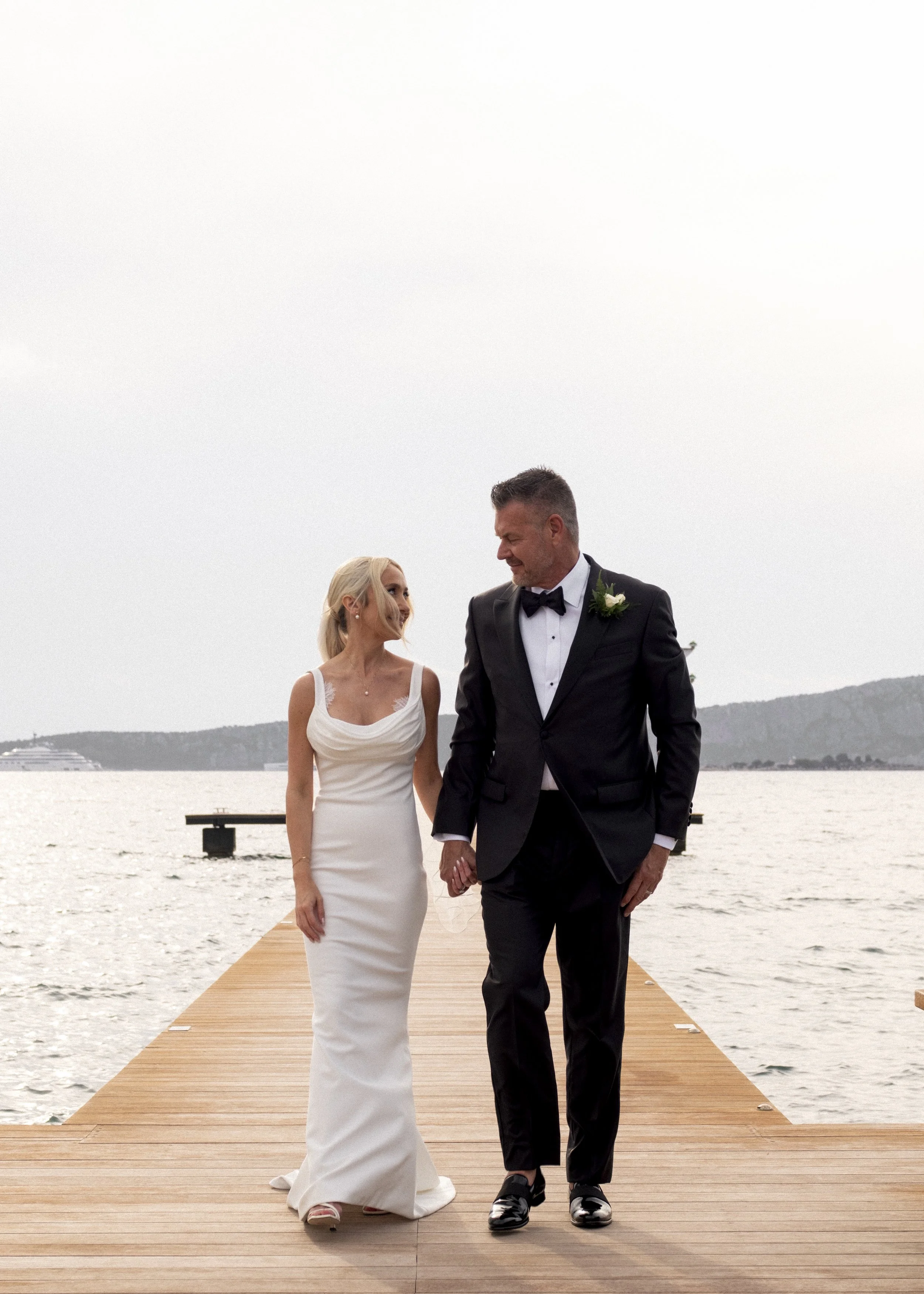 A bride and groom walking hand in hand on a wooden dock by the water, smiling and looking at each other, with hills and a cruise ship in the background during their wedding.